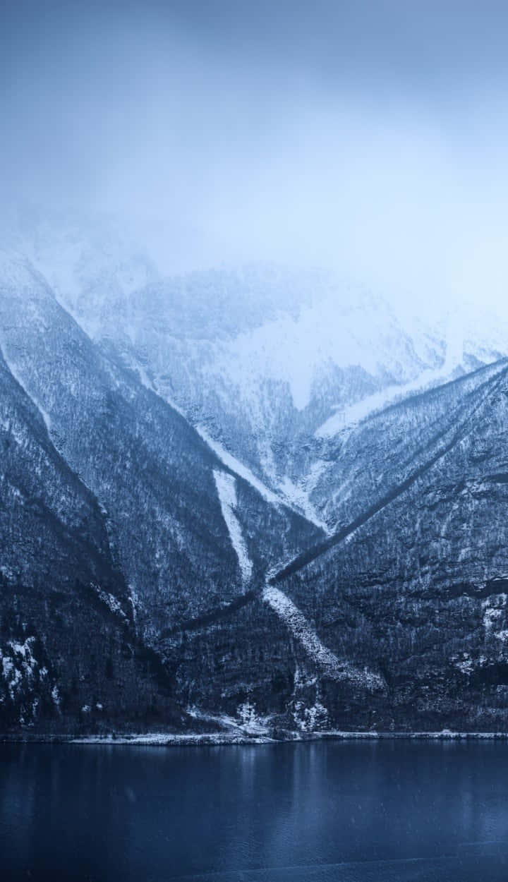 Norway 桌布 壁紙 — 一個霧氣瀰漫的冬季山谷，白雪覆蓋的山峰倒映在陰天下一片平靜的藍色湖水中。