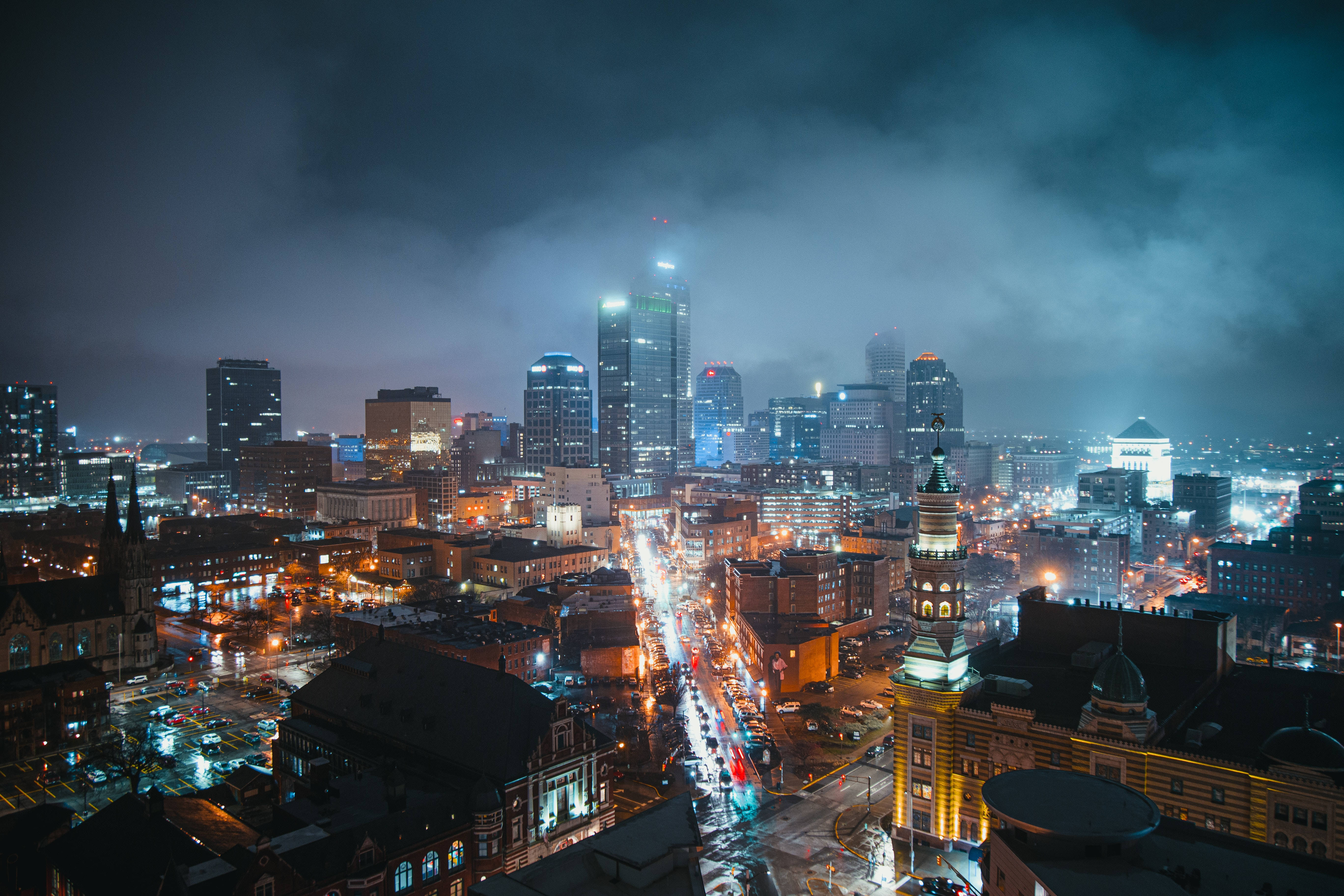 雲 桌布 壁紙 — 一個霧氣瀰漫的夜晚城市景觀，閃爍的摩天大樓和濕漉漉的街道在陰雲密布的天空下反射出鮮豔的路燈。
