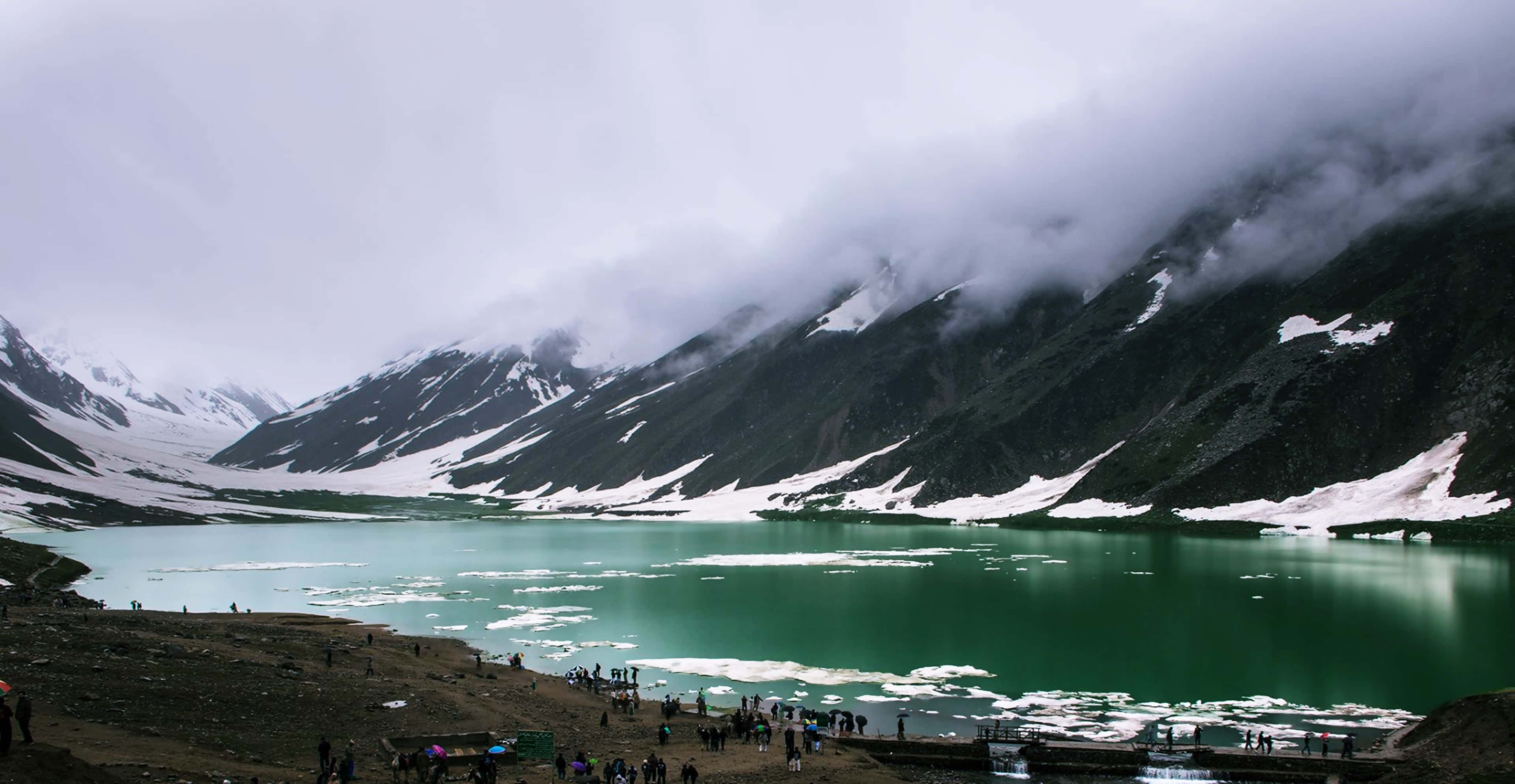 Pakistan 桌布 壁紙 — 一個霧氣瀰漫的山間湖泊，湖水呈藍綠色，山峰上覆蓋著積雪，天空多雲，人們聚集在湖岸邊。