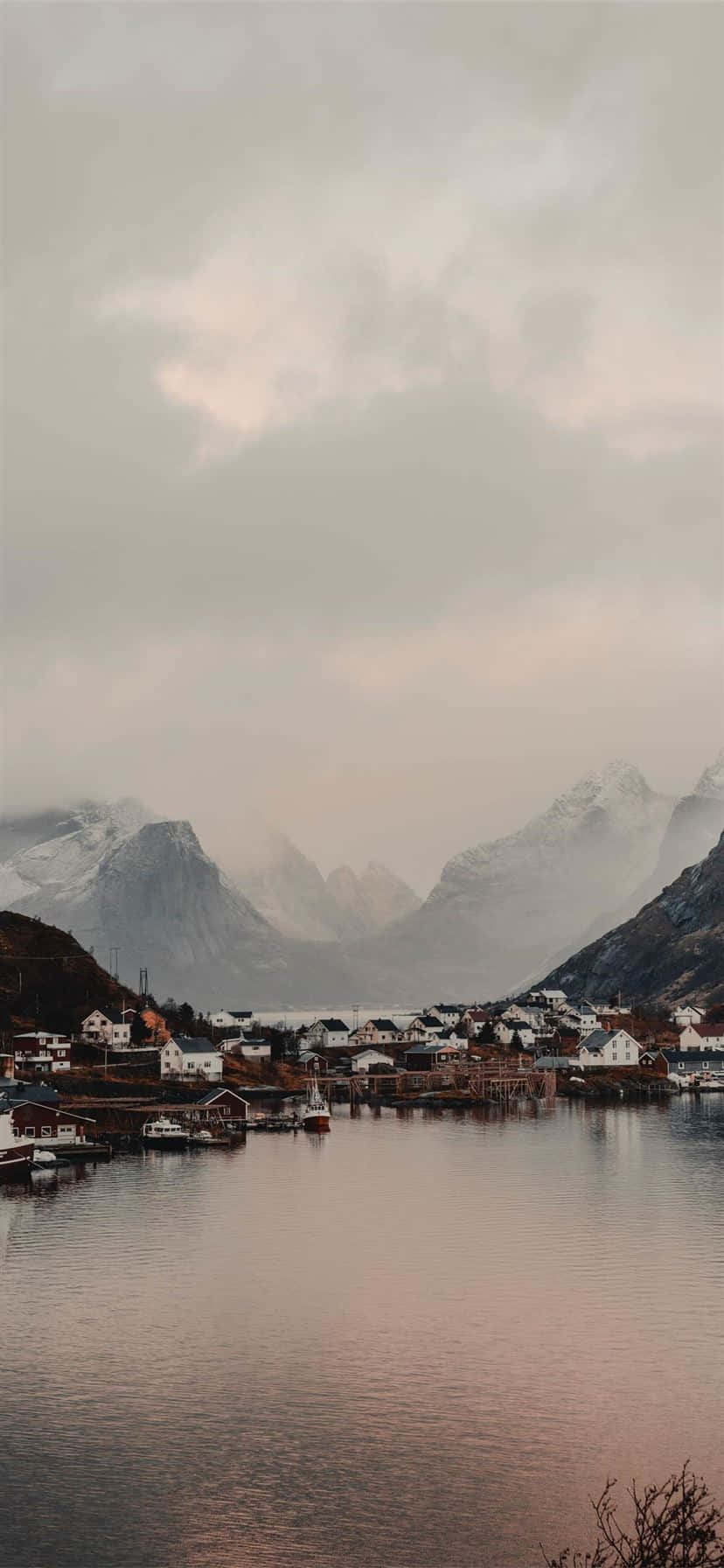 Norway 桌布 壁紙 — 一個霧氣瀰漫的峽灣村莊，背後聳立著積雪覆蓋的山脈，平靜的水面映照出柔和的粉紅色和灰色色調。