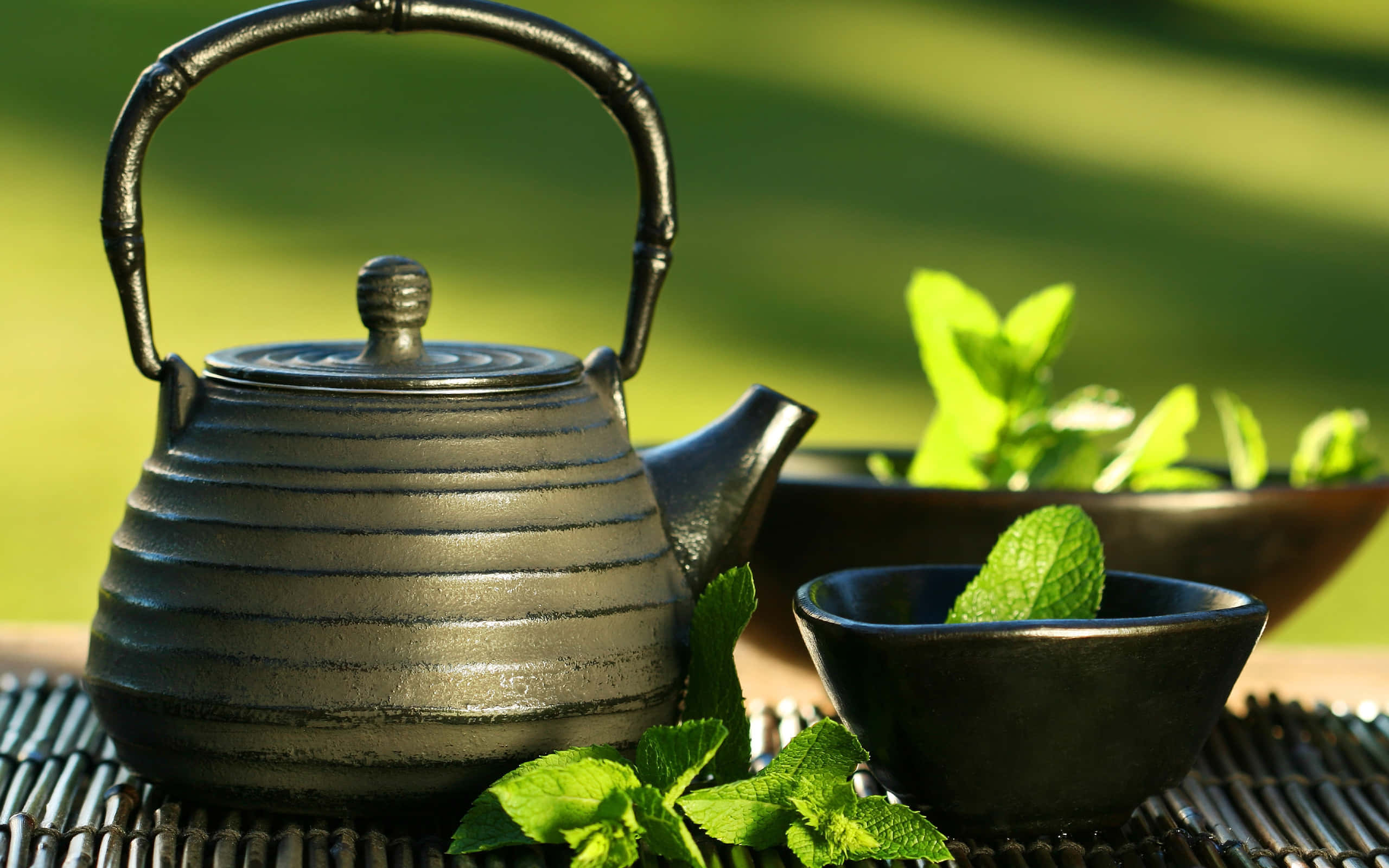 Floral Tea 桌布 壁紙 — 一個霧面黑色陶瓷茶壺和杯子放在編織墊上，旁邊是新鮮薄荷葉，背景是陽光普照的綠色草坪。