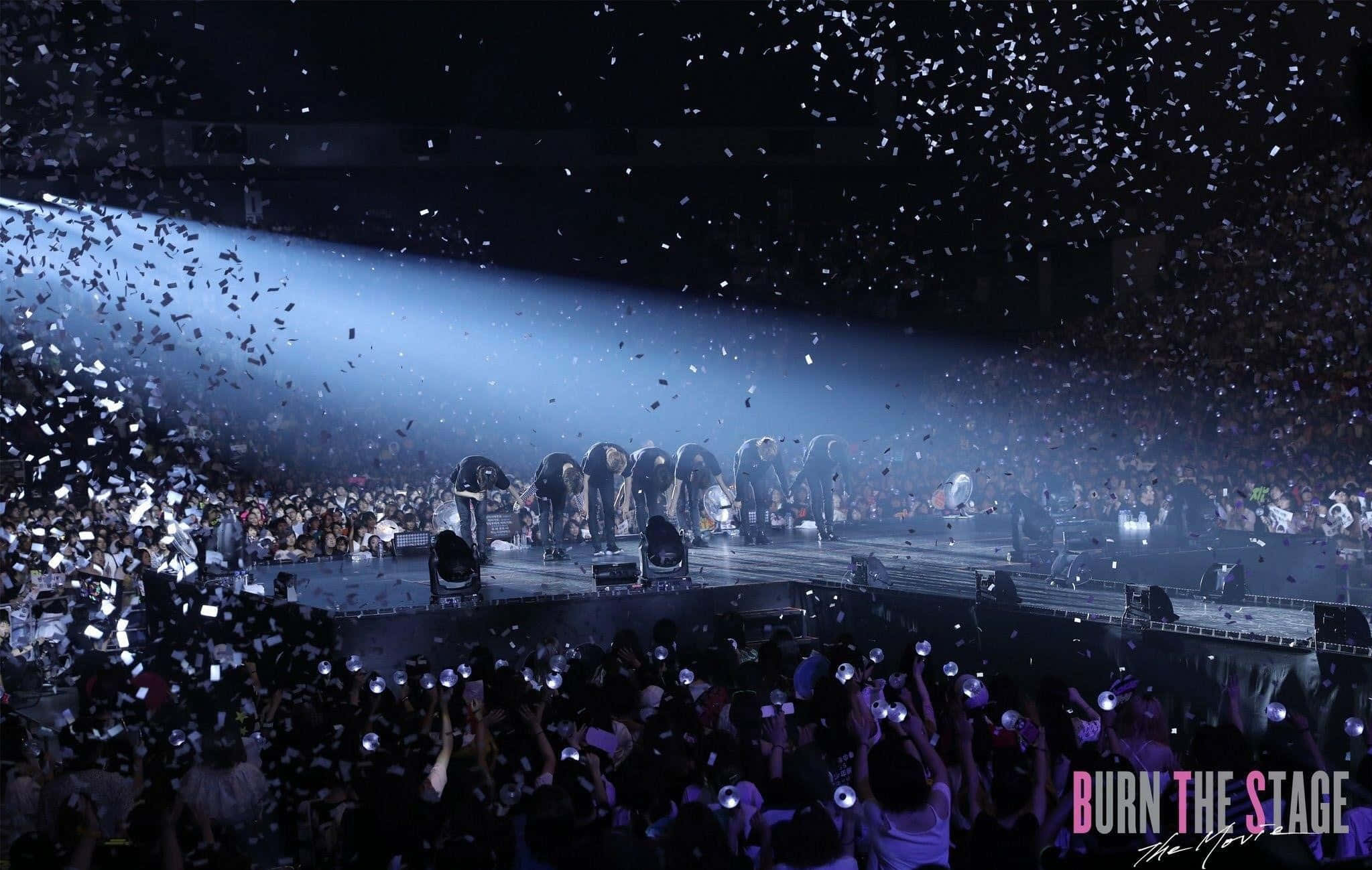 Bts Stage 桌布 壁紙 — 一個音樂會樂團在舞台上鞠躬，聚光燈下五彩紙屑如雨般灑落在高舉手機歡呼的人群上。