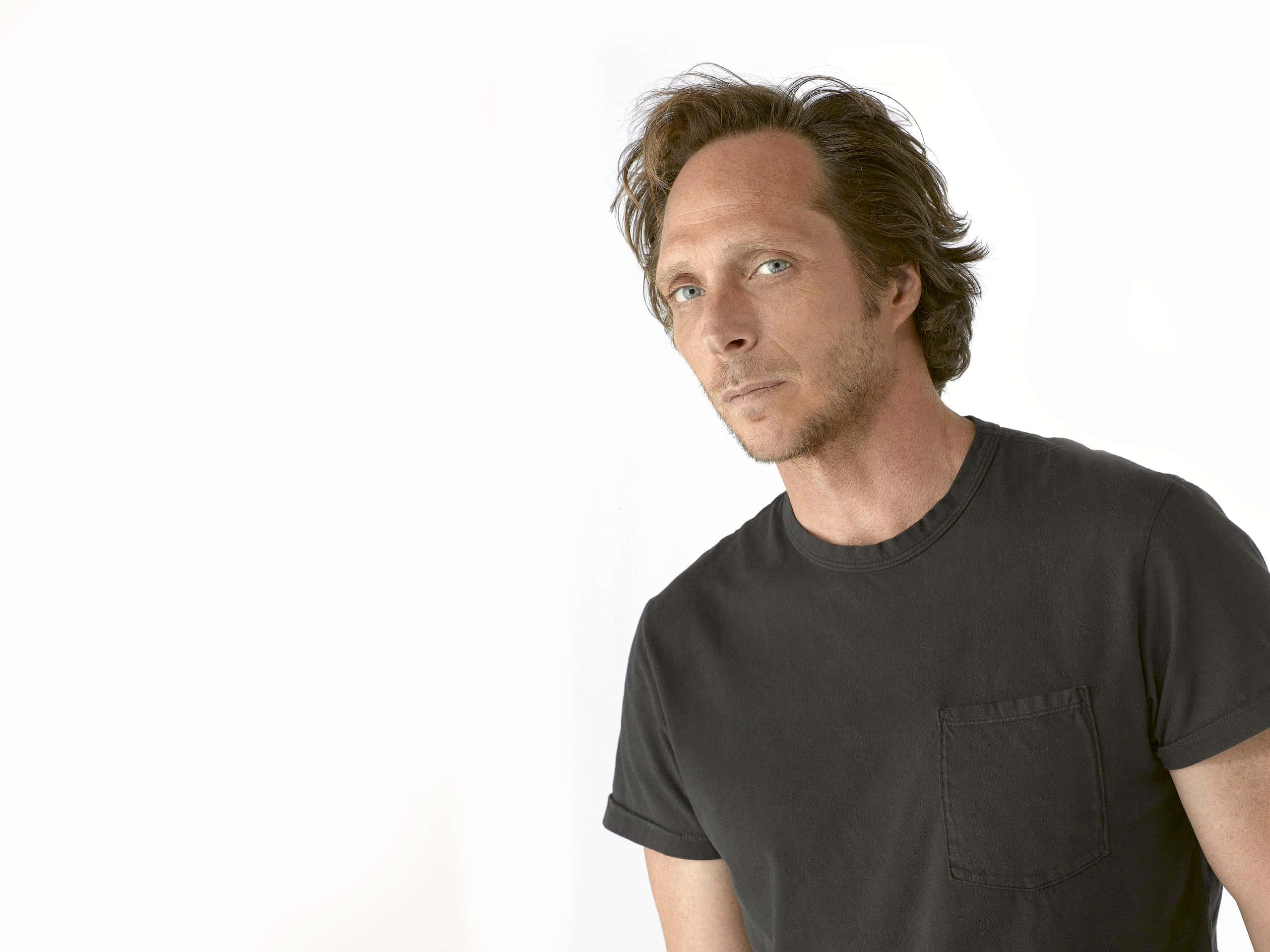 William Fichtner 桌布 壁紙 — 一個頭髮凌亂的棕色頭髮、身穿黑色T恤的男人，在純白背景下若有所思地凝視著。
