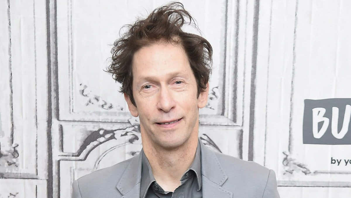 Tim Blake Nelson 桌布 壁紙 — 一個頭髮凌亂的棕色頭髮男人在白色鑲板牆前溫柔微笑，牆上有裝飾線條。