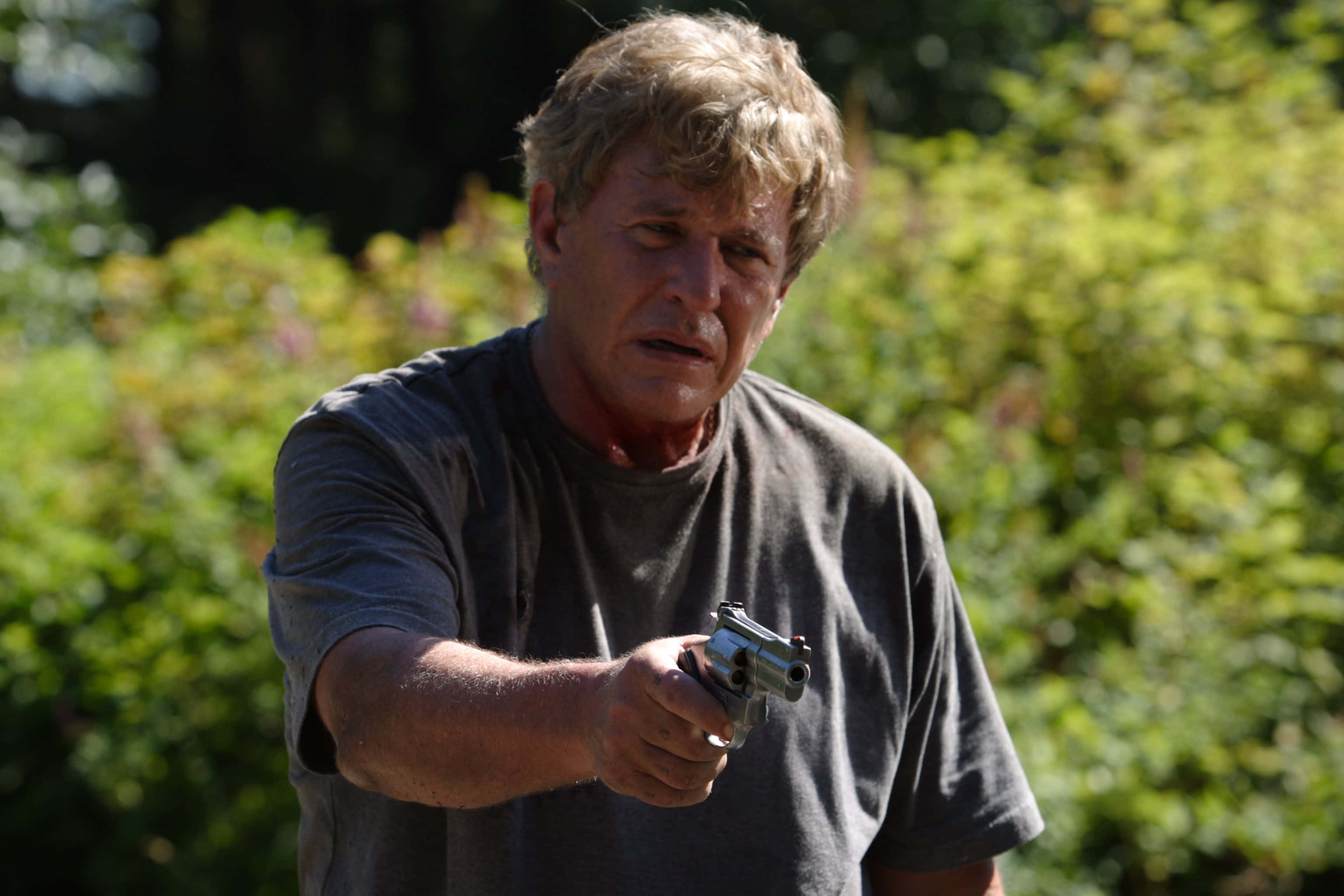 Tom Berenger 桌布 壁紙 — 一個頭髮凌亂的男人，在陽光照射的綠色灌木背景下，表情緊張地向前瞄準一把銀色手槍。