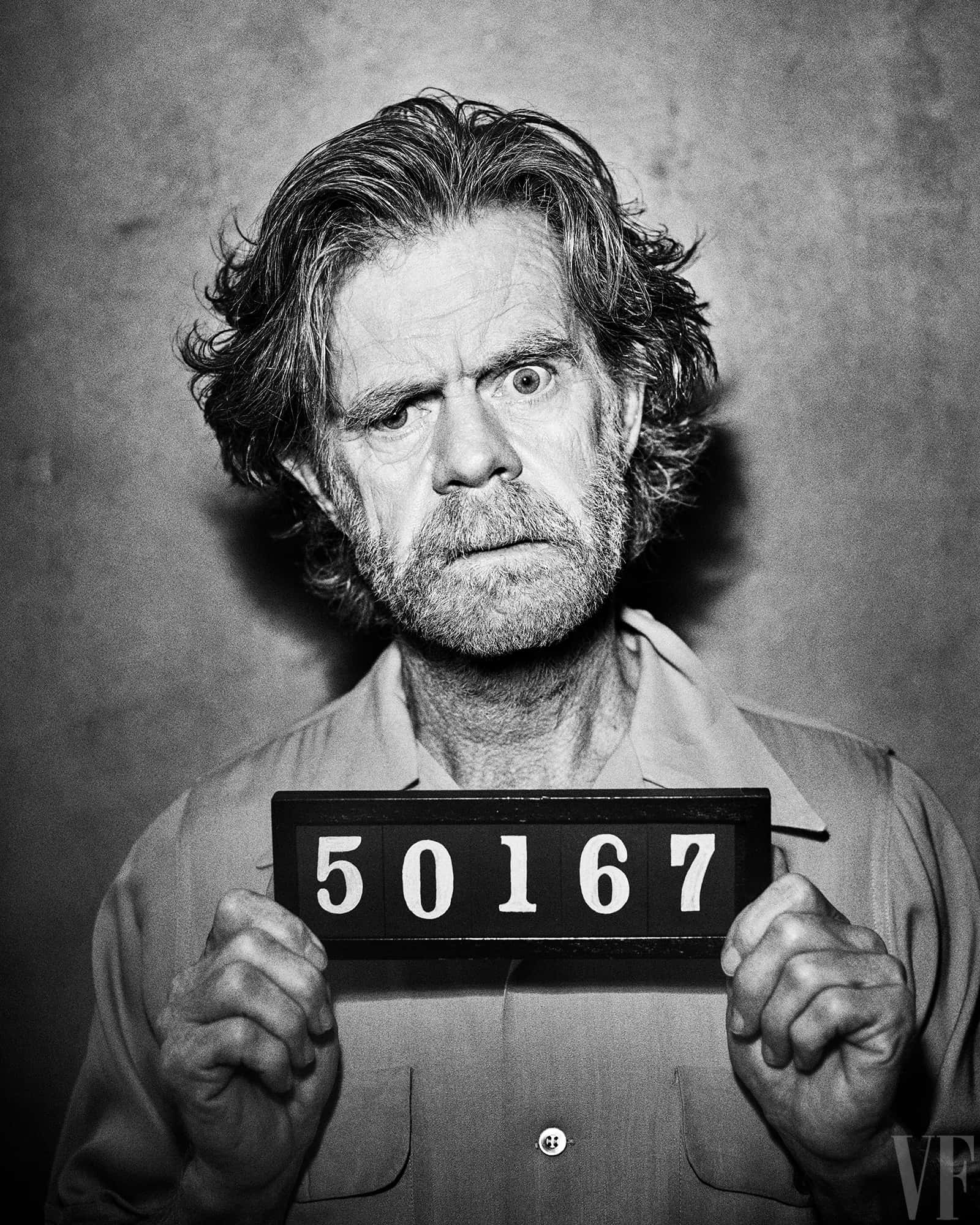 william h. macy 桌布 壁紙 — 一個頭髮狂野、眼神銳利的男人，在清晰的黑白肖像中拿著一張 mugshot 號碼牌。