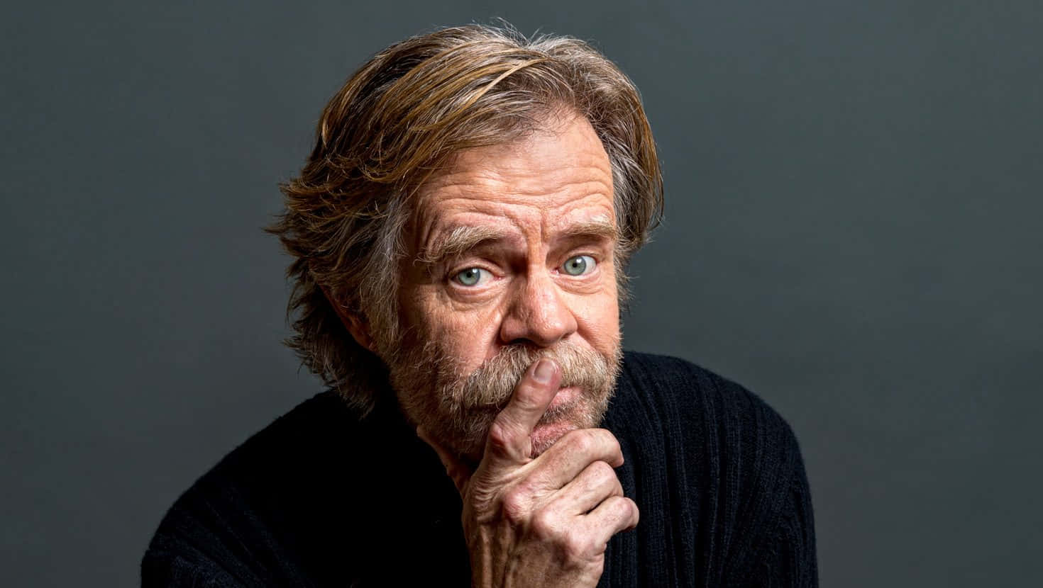william h. macy 桌布 壁紙 — 一個頭髮花白、留鬍鬚的男人，在深灰色背景前，將手指放在嘴唇上，專注地凝視著。