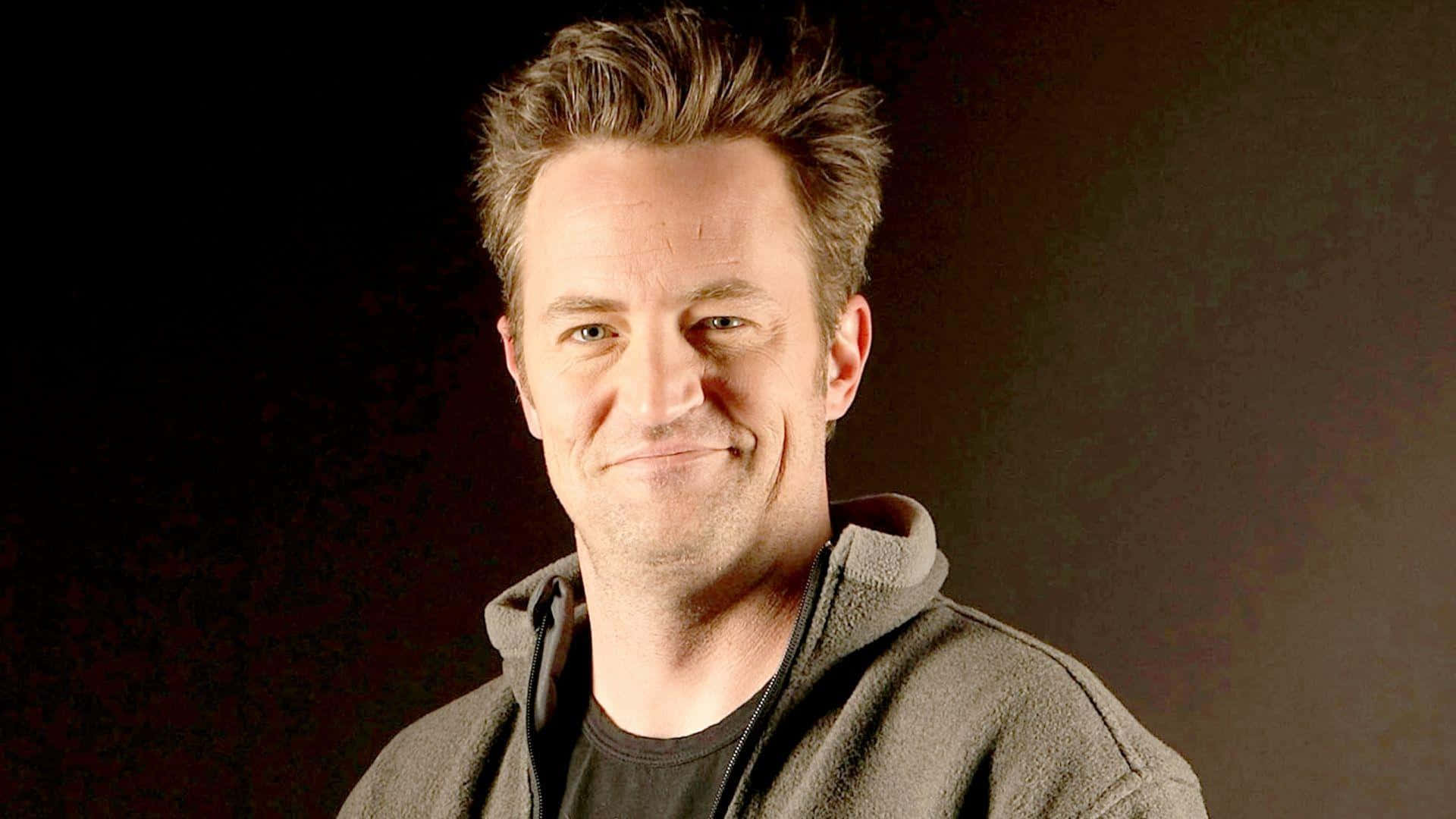 matthew perry 桌布 壁紙 — 一個頭髮蓬鬆的男人穿著深色連帽衫，在黑色和棕色漸變背景前溫暖地微笑。