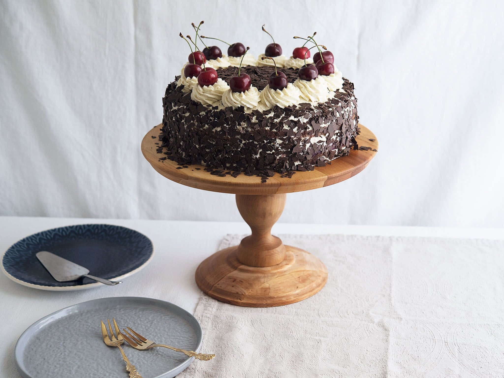 Black Forest Cake 桌布 壁紙 — 一個頹廢的黑森林蛋糕，飾以鮮奶油和光澤櫻桃，優雅地放在木架上，襯以柔和的白色背景。