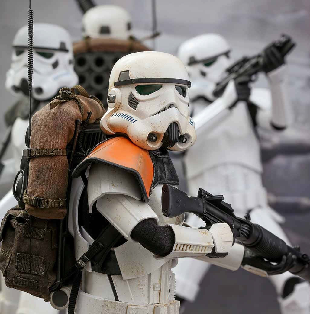 Jedha 桌布 壁紙 — 一個風化嘅暴風兵公仔嘅特寫，佢着住橙色背心同揹住背囊，揸住一支爆能槍，周圍有其他士兵喺一個粗糙嘅場景入面。
