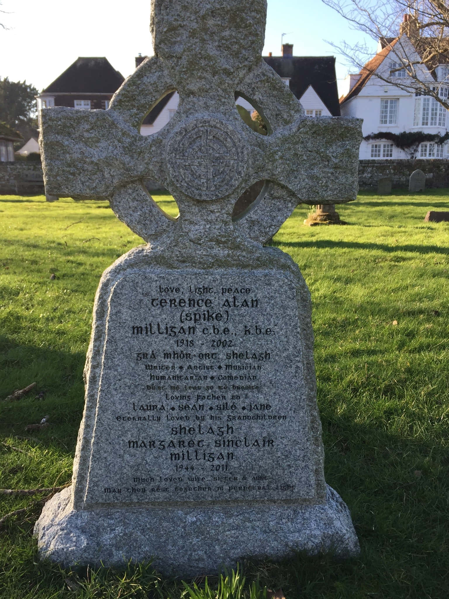Gravestone 桌布 壁紙 — 一個風化的凱爾特十字墓碑，位於陽光明媚的墓地，有修剪整齊的草地和背景中的房屋。
