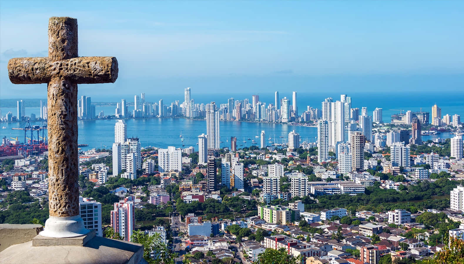 Colombia 桌布 壁紙 — 一個風化的石十字架俯瞰著廣闊的海岸城市景觀，現代摩天大樓和港口在清澈的藍天下。