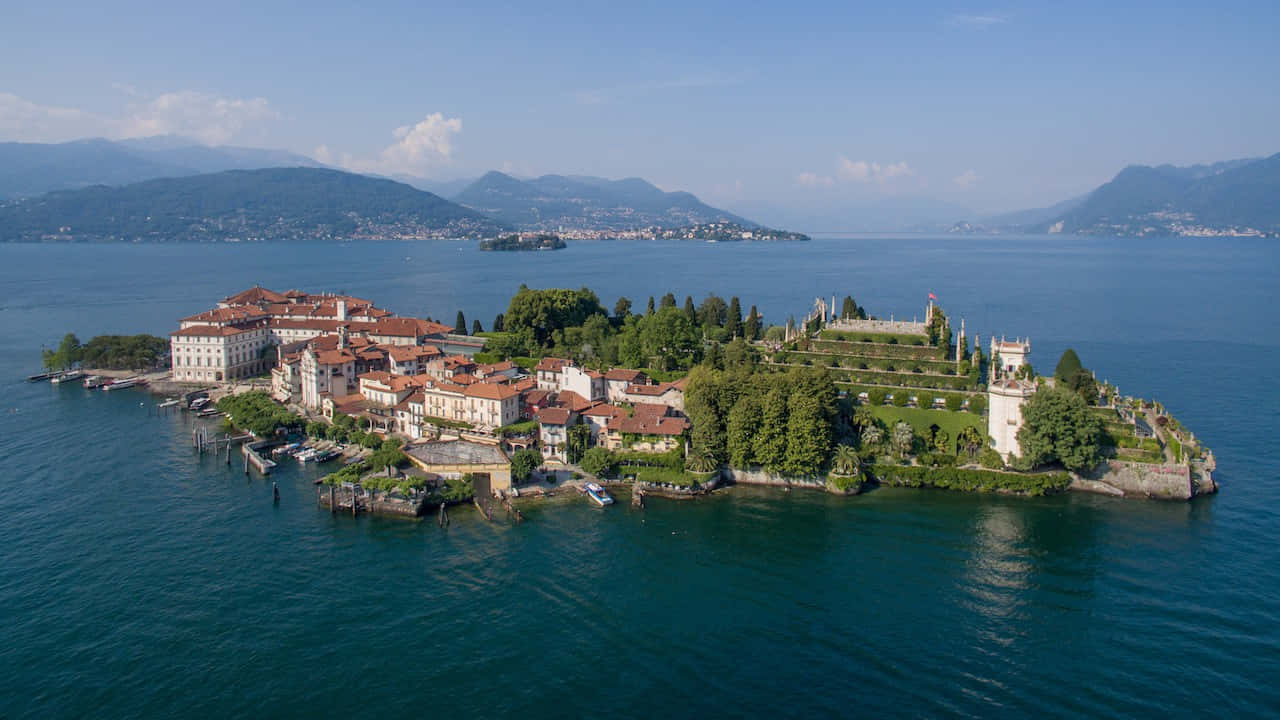 Italian Island 桌布 壁紙 — 一個風景如畫的島嶼的鳥瞰圖，島上有赤陶屋頂的建築物和茂密的花園，周圍是深藍色的海水和遠處的山脈，天空晴朗。