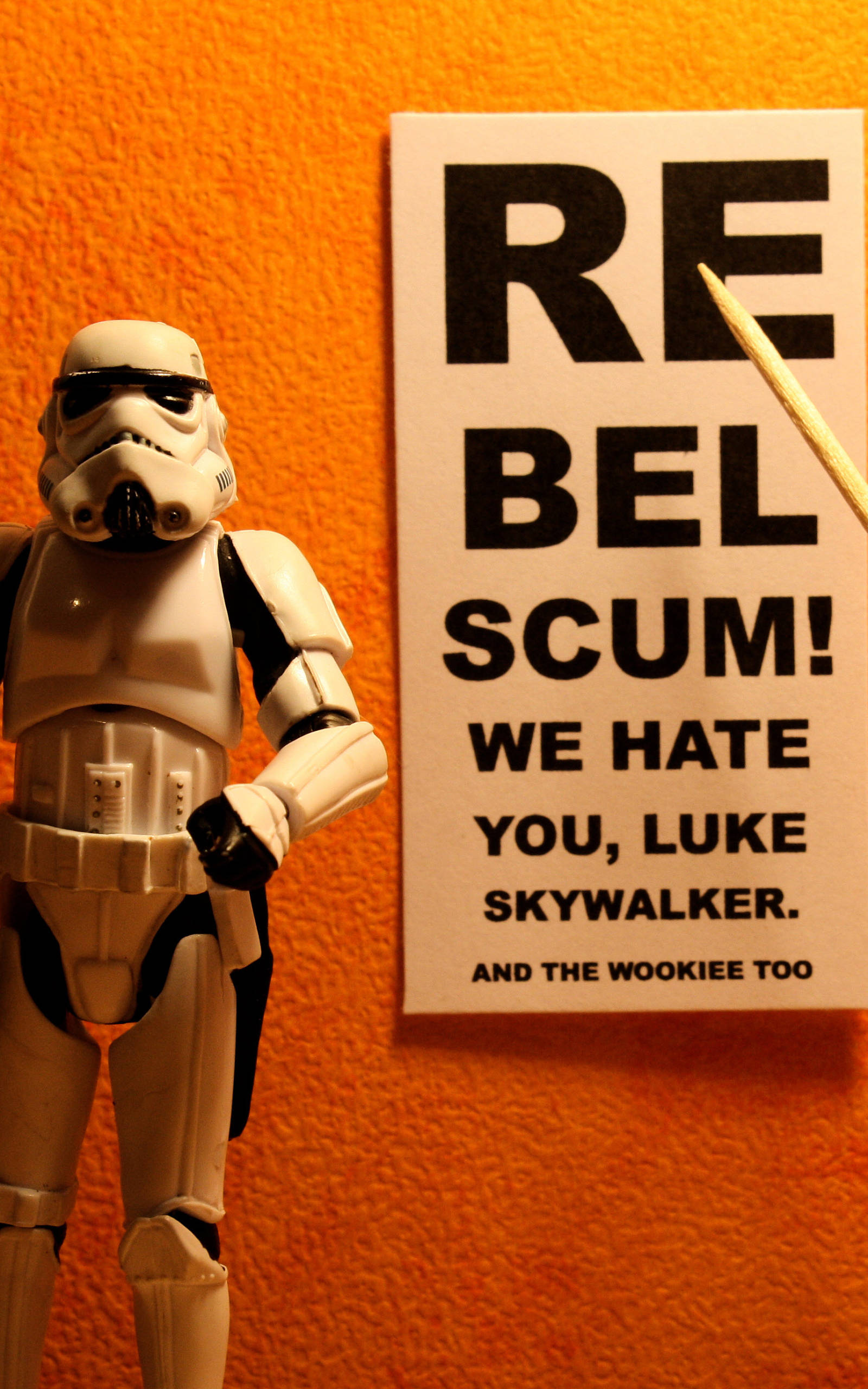 星際大戰 桌布 壁紙 — 一個風暴兵可動人偶站在一個寫著 rebel scum we hate you luke skywalker and the wookiee too 的標誌旁邊，背景是橙色牆壁。