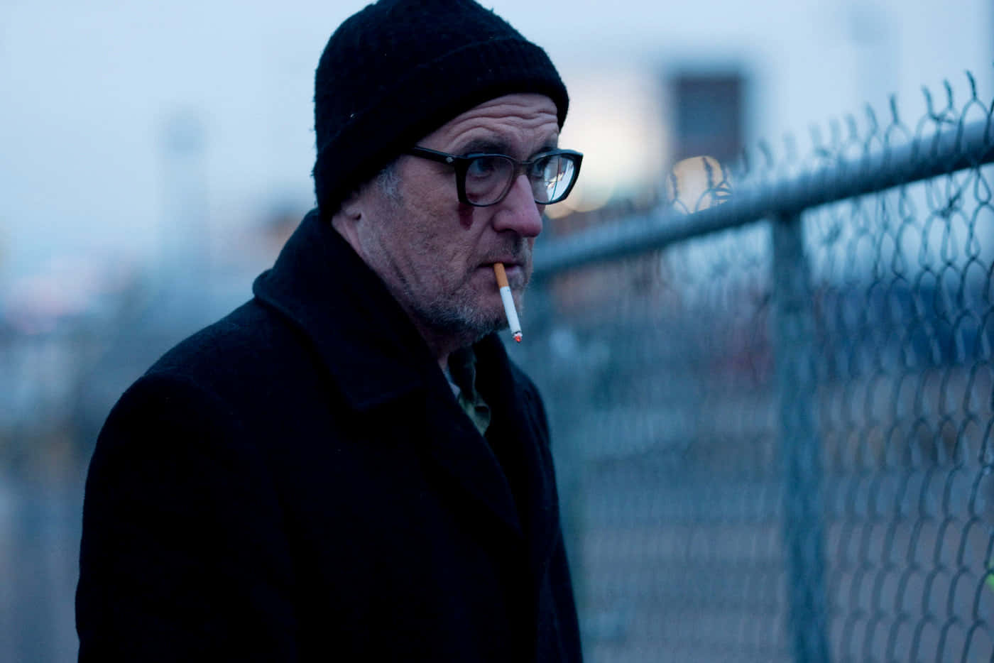 Richard Jenkins 桌布 壁紙 — 一個飽經風霜的男人戴着黑色冷帽和外套，在陰沉天空下的鐵絲網旁抽煙。