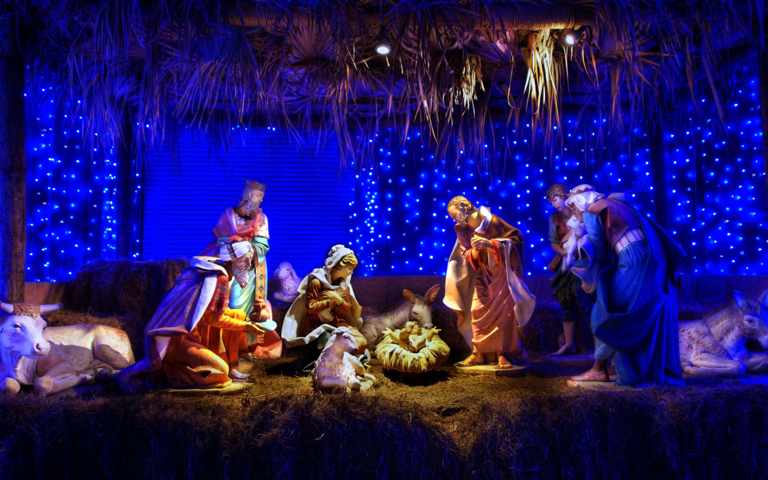 Nativity 桌布 壁紙 — 一個馬槽場景，人物雕像在茅草屋頂下，由藍色仙女燈和溫暖的聚光燈照亮。