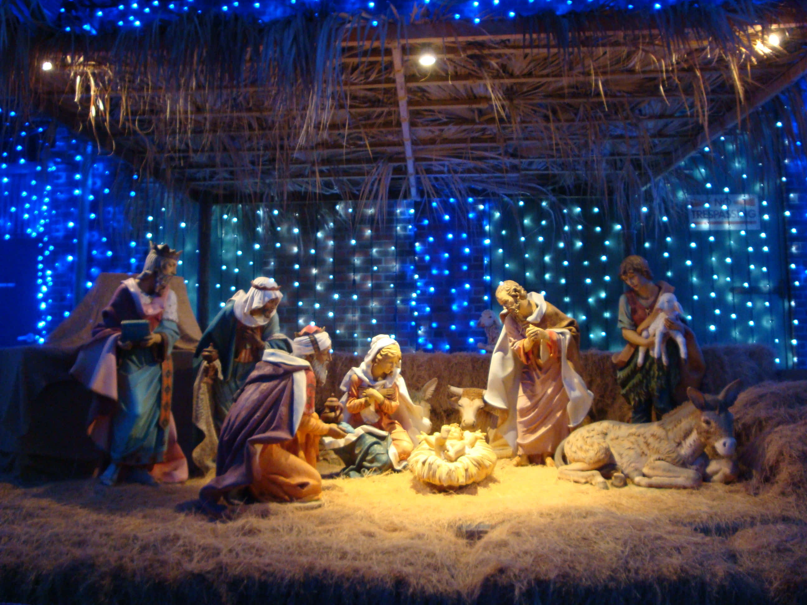 Nativity 桌布 壁紙 — 一個馬槽場景，有瑪利亞、約瑟、嬰孩耶穌、牧羊人和動物的雕像，在茅草屋頂下，由藍色燈串照亮。