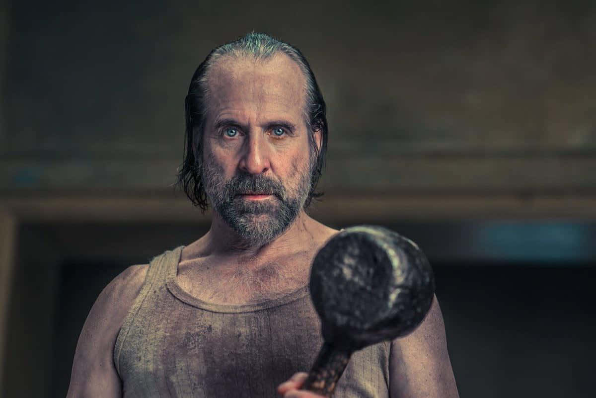Peter Stormare 桌布 壁紙 — 一個骯髒鬍鬚男，身穿污跡背心，在昏暗房間內緊握重錘，眼神凌厲。