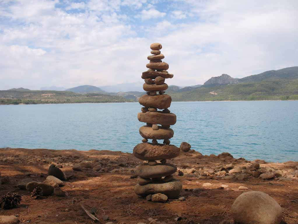 Land Art 桌布 壁紙 — 一個高大的平衡石堆矗立在乾燥的湖岸上，平靜的藍色水域和遠處的綠色山丘在部分多雲的天空下。