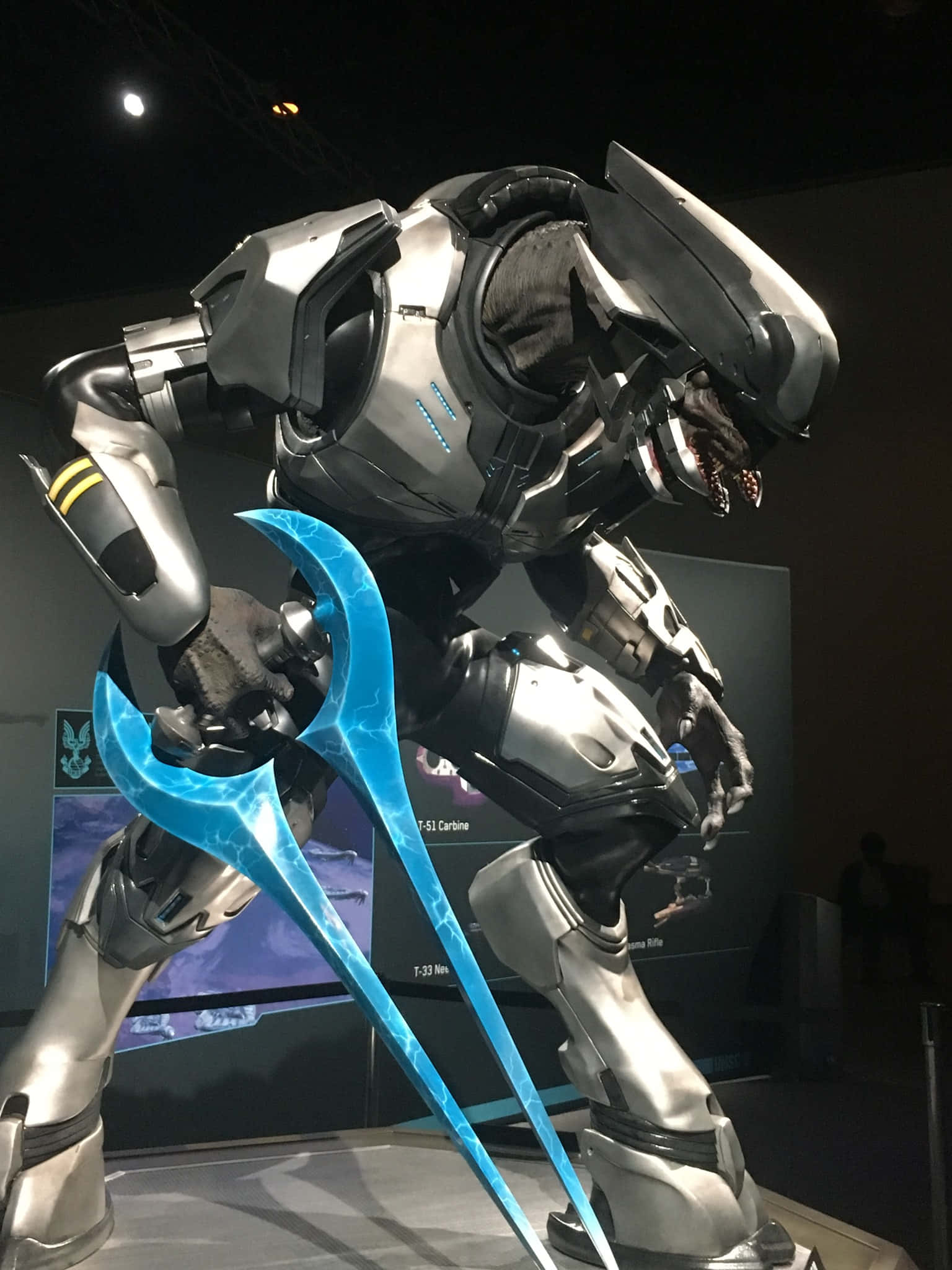 Sangheili 桌布 壁紙 — 一個高大的銀色裝甲戰士，手持發光的藍色能量武器，站在燈光昏暗的展覽廳中。