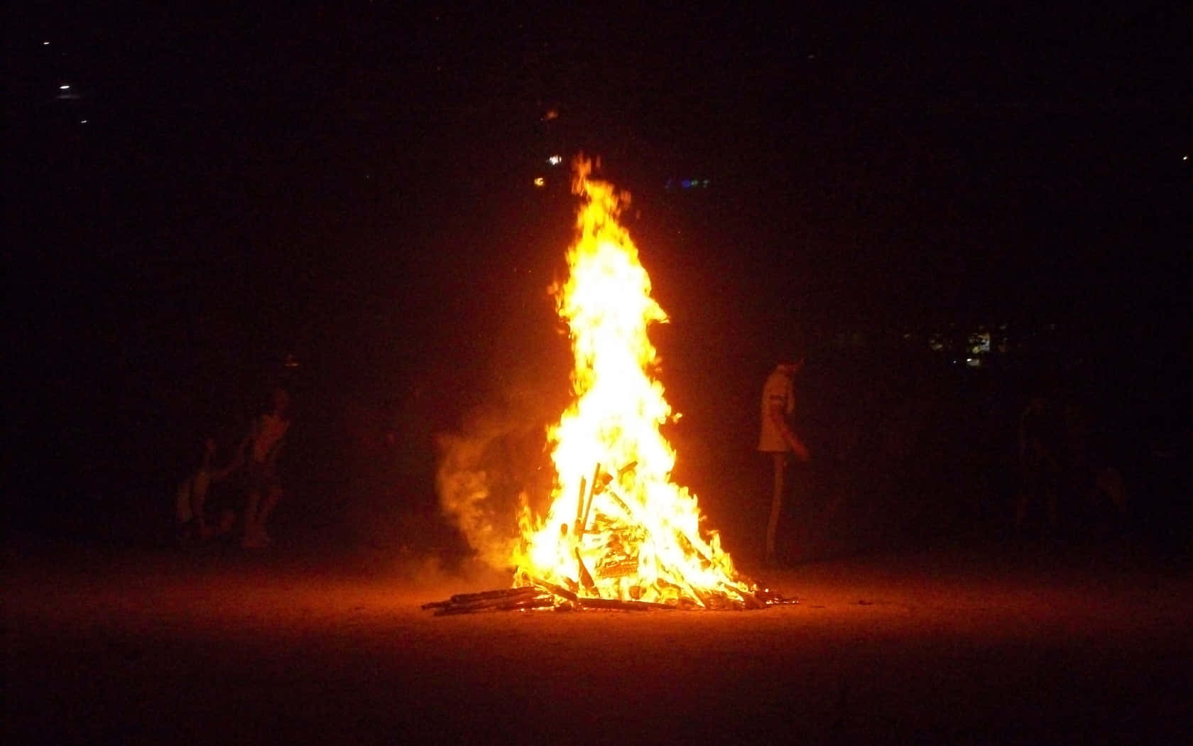 Fall Bonfire 桌布 壁紙 — 一個高聳的營火在漆黑的夜空下猛烈燃燒，發出明亮的橙色火焰，周圍聚集著剪影般的人影。