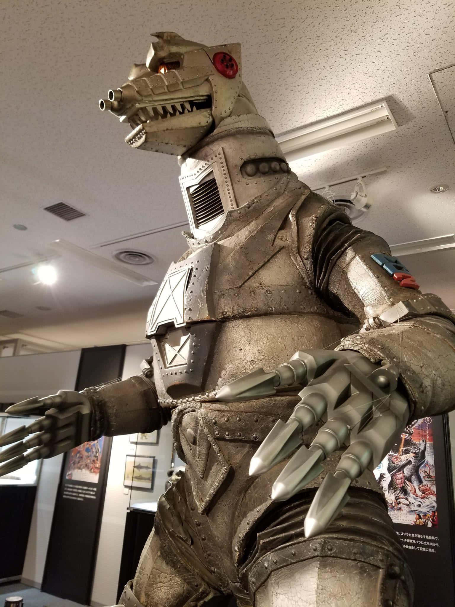 mechagodzilla 桌布 壁紙 — 一個高聳的金屬機械怪物，帶有尖刺爪子和發光的紅色眼睛，在明亮的燈光下站在博物館展覽中。