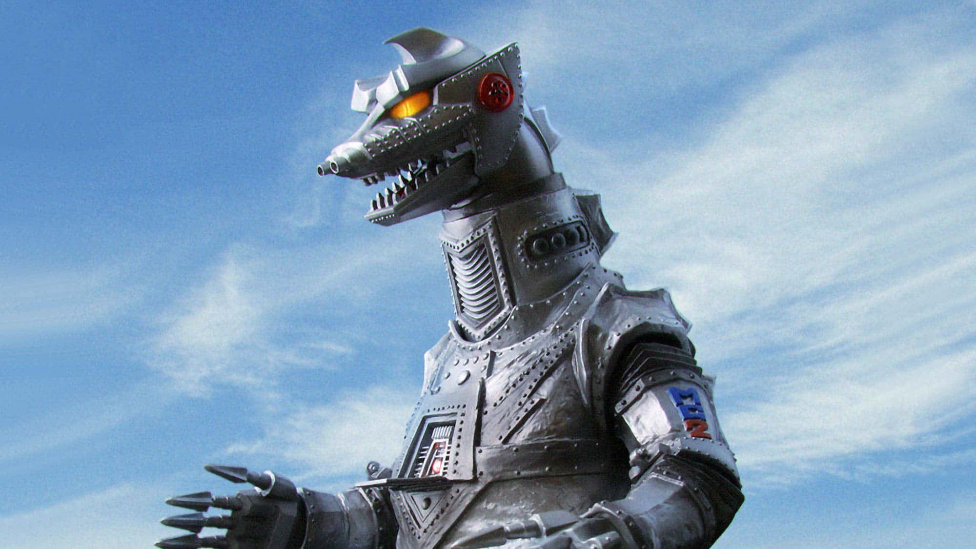 Robot Godzilla 桌布 壁紙 — 一個高聳的銀色機械人怪物，擁有發光的橙色眼睛和鋒利的牙齒，站在有縷縷白雲的明亮藍天下。