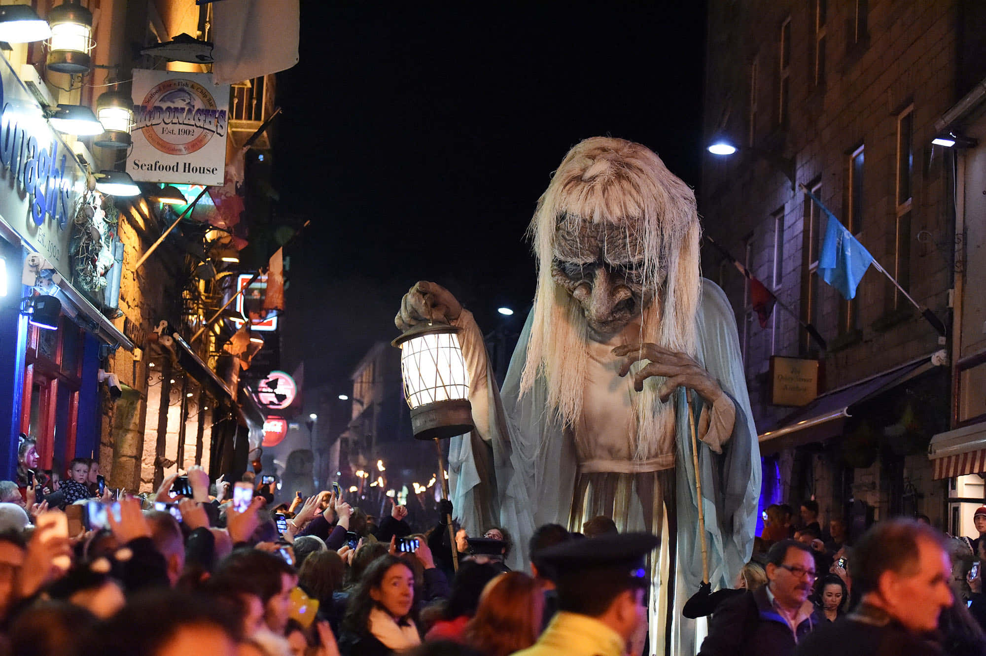 Halloween Parades 桌布 壁紙 — 一個高聳的鬼魅身影，蒼白的頭髮和提燈，穿過擁擠的夜晚街道，旁觀者拍攝這一奇觀。