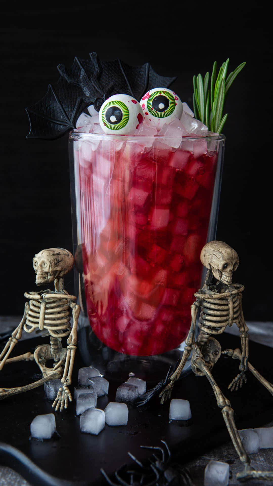 Spooky Cocktails 桌布 壁紙 — 一個高身玻璃杯裝著紅色雞尾酒，加冰塊，上面放著彈珠眼、蝙蝠翼和迷迭香，周圍有細小骷髏，放在深色表面上。