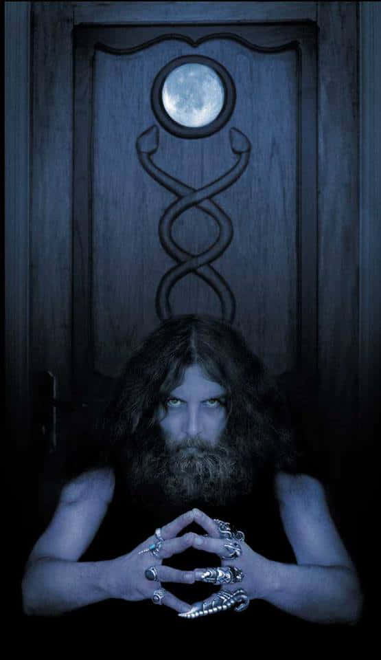 Alan Moore 桌布 壁紙 — 一個鬍鬚男，眼睛發光，戴著華麗戒指，透過一扇帶有月亮和扭曲結設計的深色木門凝視。