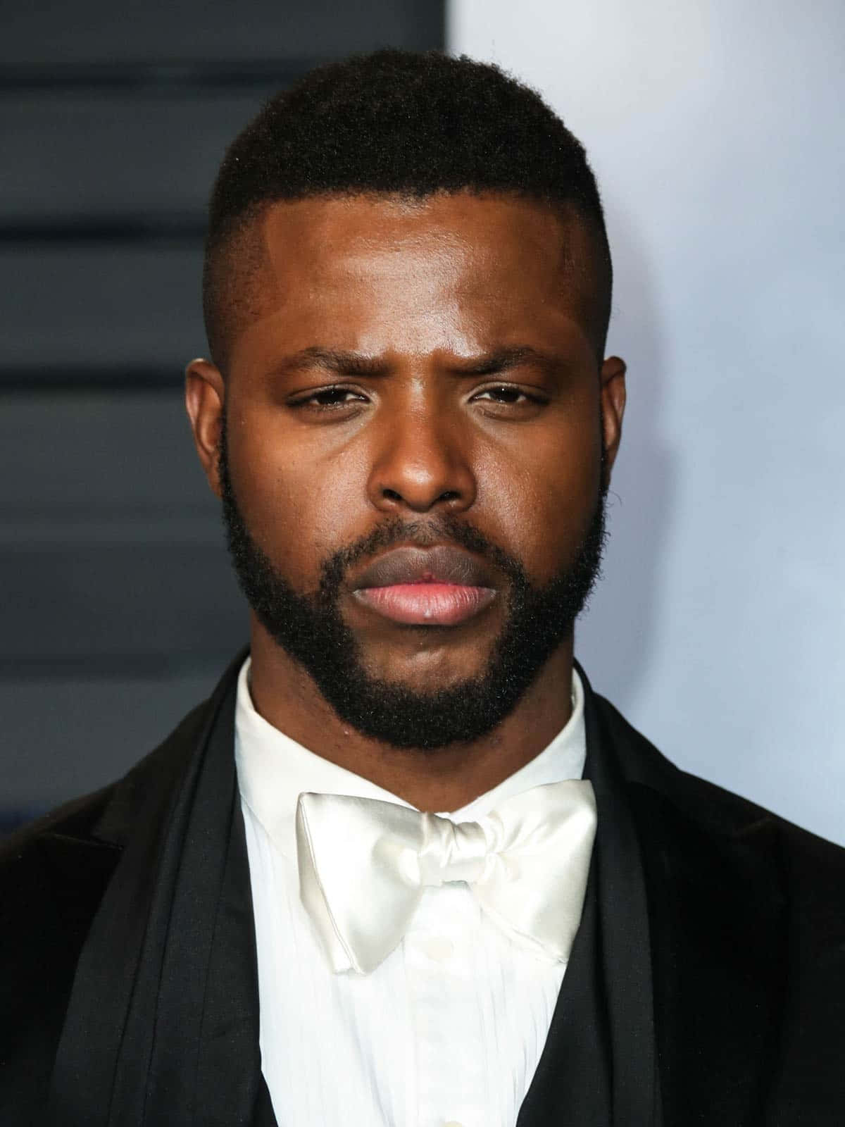 Winston Duke 桌布 壁紙 — 一個鬍鬚男人的特寫肖像，穿著黑色燕尾服和白色領結，背景模糊。