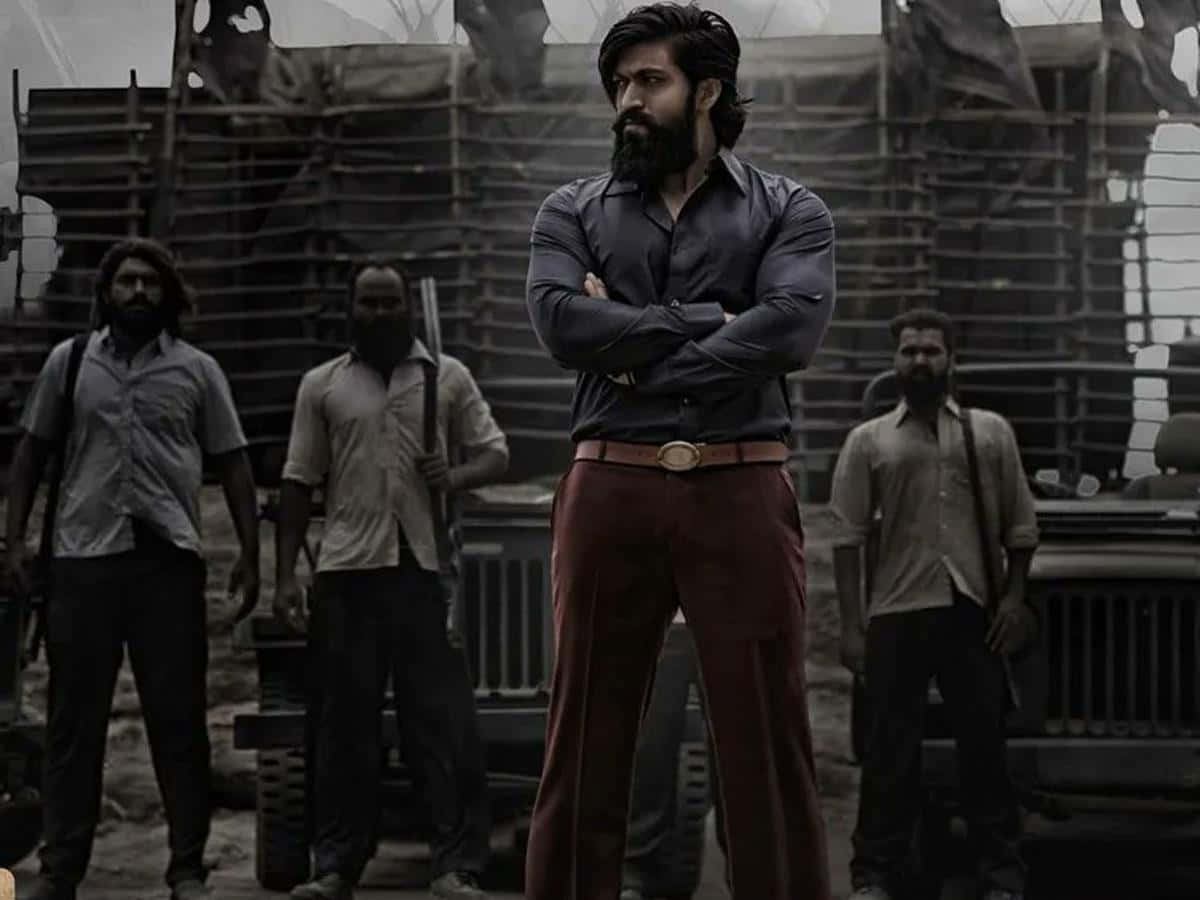 Kgf 2 桌布 壁紙 — 一個鬍鬚男人穿著深色襯衫和紅色褲子，自信地交叉雙臂站立，被四個武裝男子夾在一個粗糙的工業環境中。