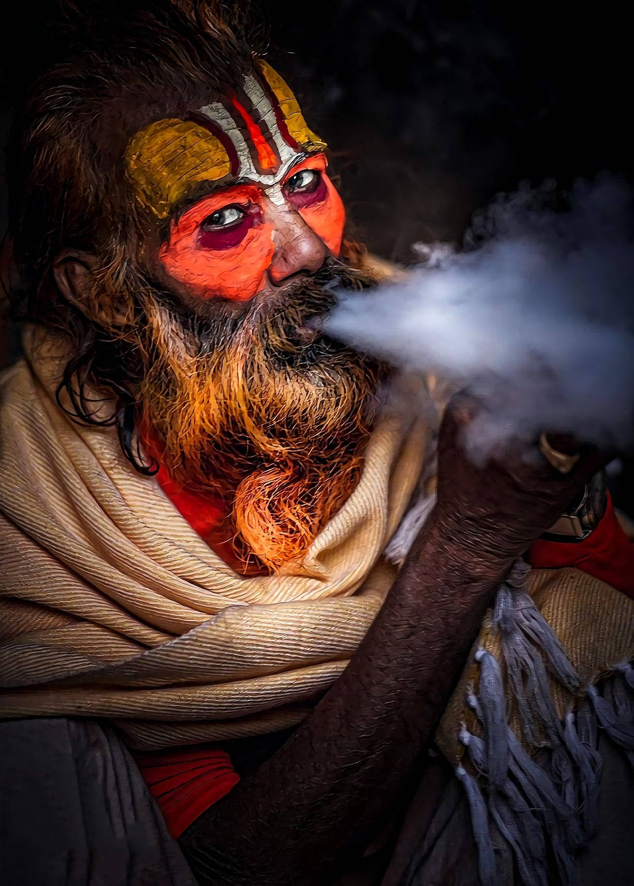 Aghori 桌布 壁紙 — 一個鬍鬚男嘅特寫，佢面上有鮮艷嘅橙色同金色顏料，喺黑暗背景前呼出煙霧。