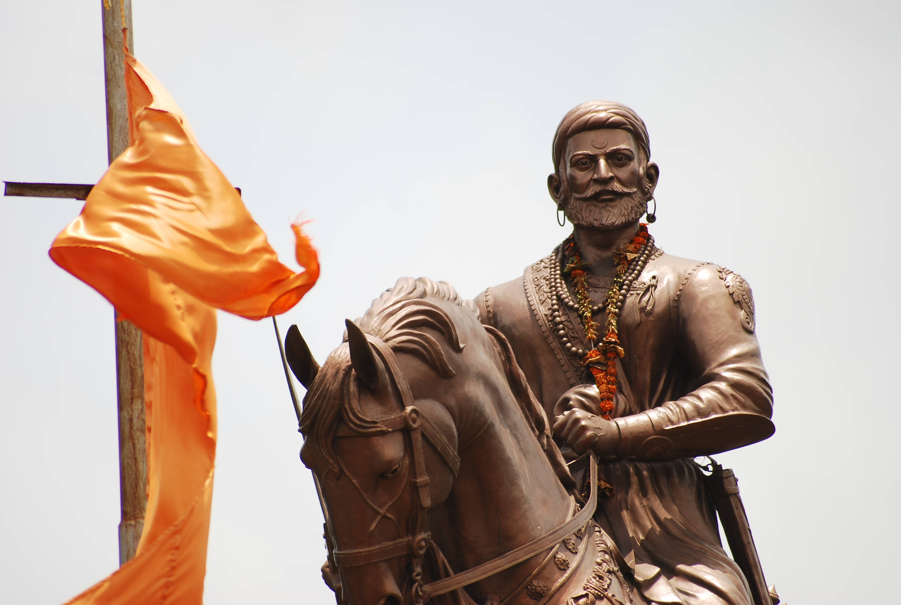 Indian 桌布 壁紙 — 一個鬍鬚男子的青銅騎馬雕像，身穿傳統服飾，飾有橙色花環和珠寶，旁邊有一面鮮豔的橙色旗幟在蒼白的天空下飄揚。
