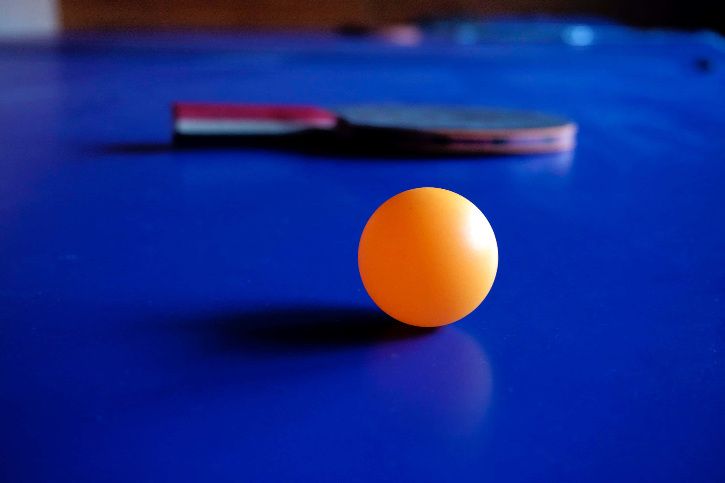 ball 桌布 壁紙 — 一個鮮橙色嘅乒乓球清晰聚焦喺深藍色嘅枱上，背景有塊模糊嘅球拍。