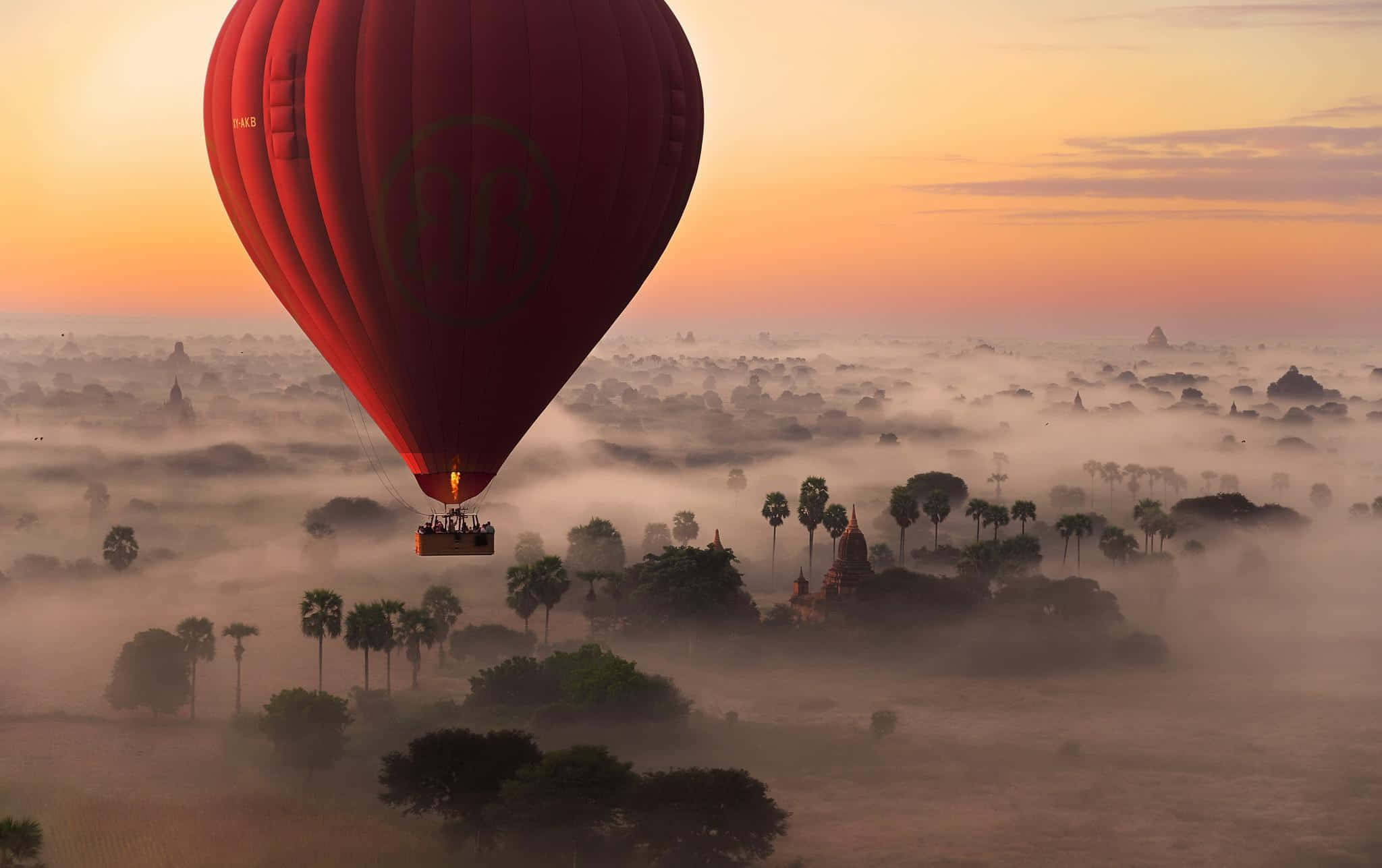 熱氣球 桌布 壁紙 — 一個鮮紅色的熱氣球漂浮在霧氣瀰漫的風景之上，點綴著古老的寺廟和棕櫚樹，正值日出時分。