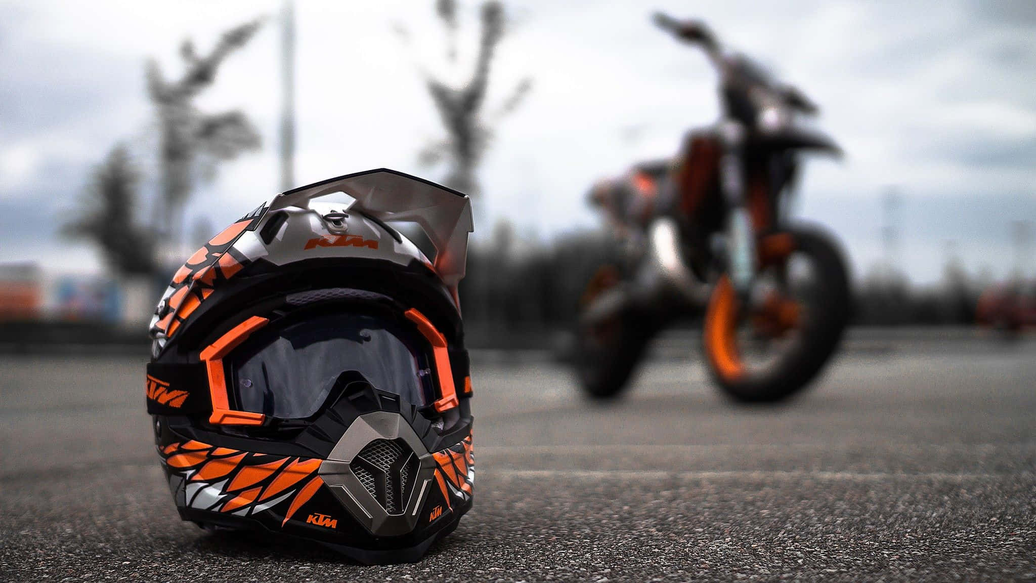 Ktm Bike 桌布 壁紙 — 一個鮮豔的橙色和黑色KTM越野電單車頭盔放在瀝青路上，背景有一輛模糊的越野電單車，天空多雲。