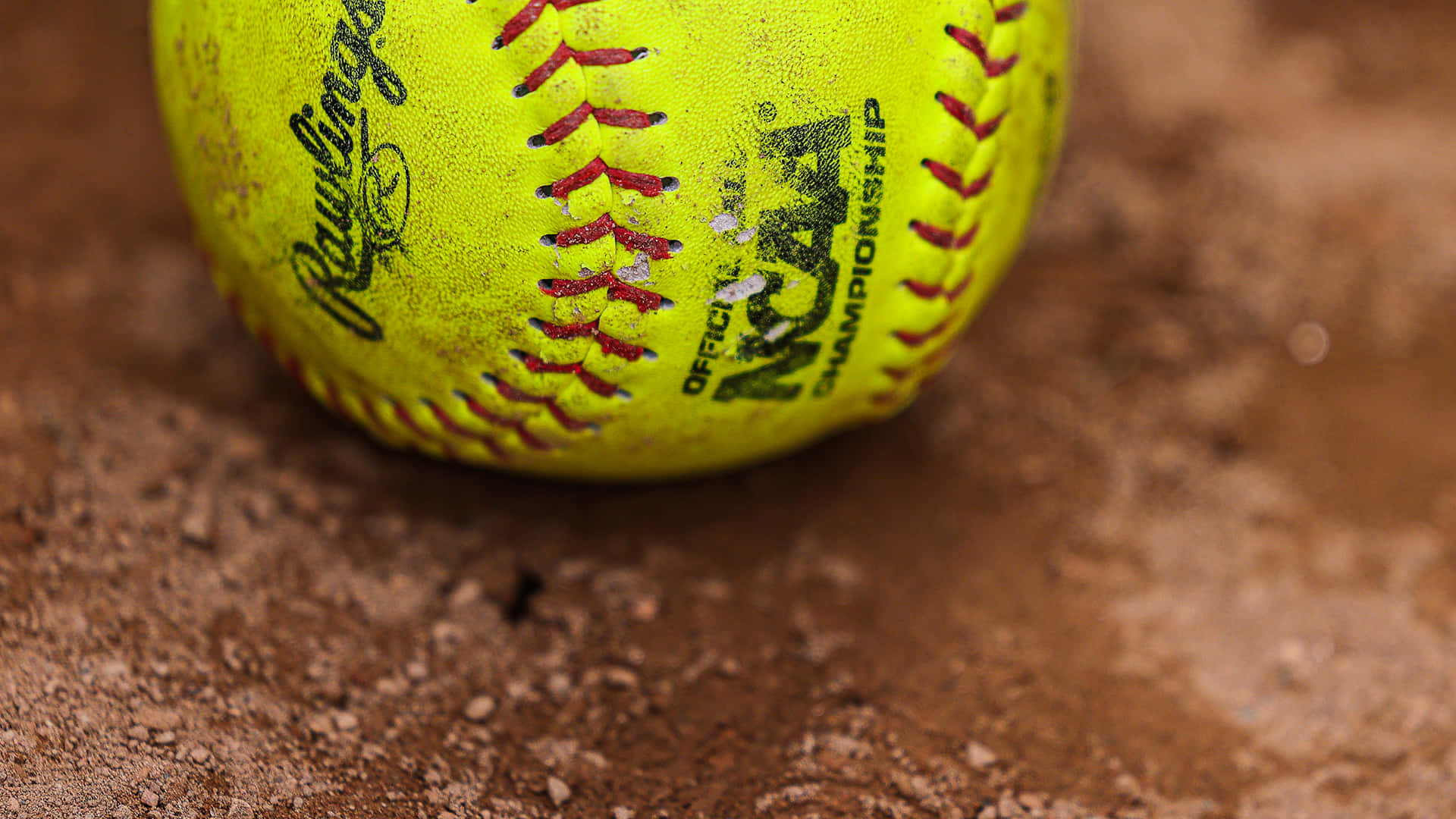 Softball 桌布 壁紙 — 一個鮮黃色NCAA壘球嘅特寫，有紅色縫線，放喺紅褐色泥土上面。