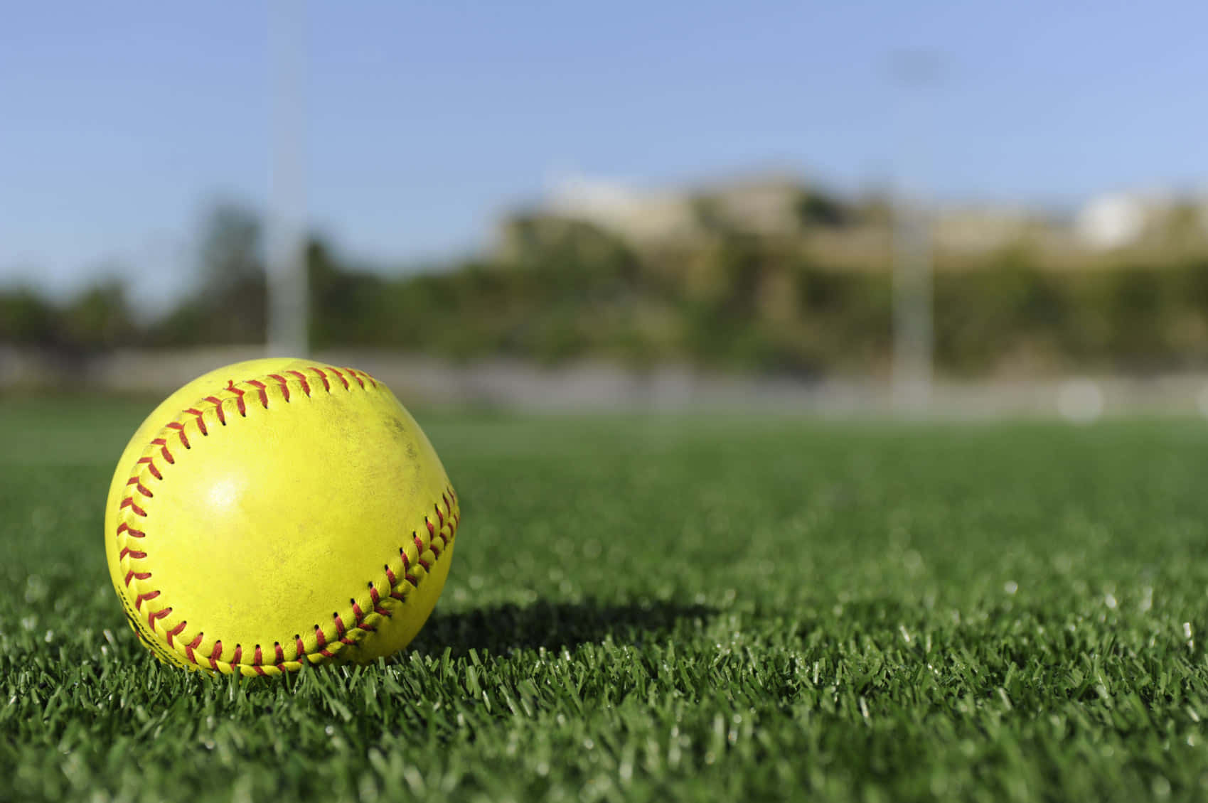 Softball 桌布 壁紙 — 一個鮮黃色嘅壘球，帶有紅色縫線，喺晴朗藍天下放喺充滿活力嘅綠色人造草皮上。
