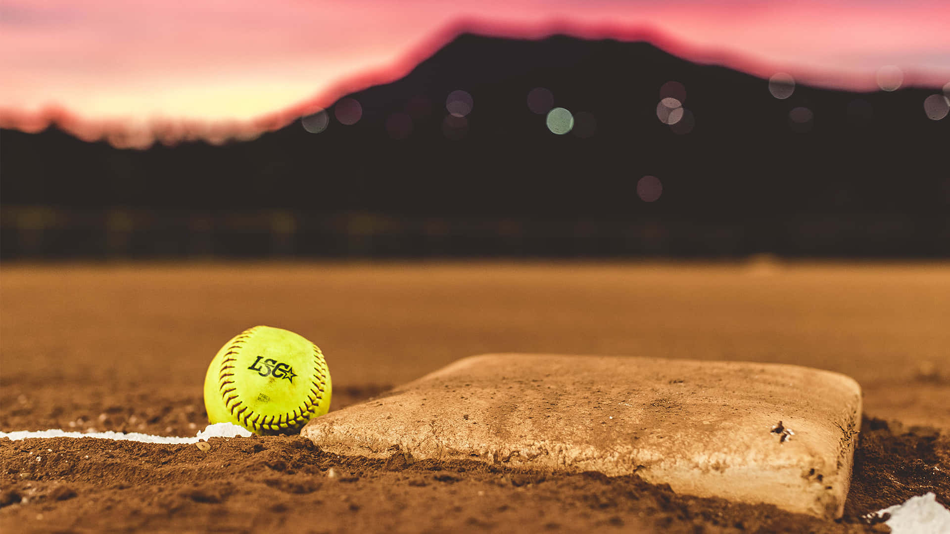 Softball 桌布 壁紙 — 一個鮮黃色的壘球放在日落時分泥土場地上磨損的本壘板旁邊，背景有山脈。