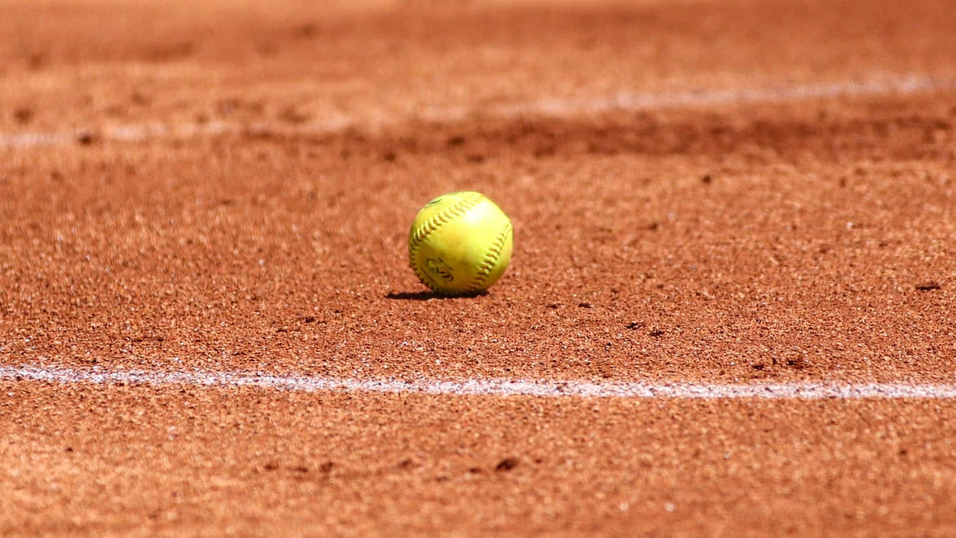 Softball 桌布 壁紙 — 一個鮮黃色的壘球放在陽光普照的紅土球場上，球場有清晰的白色界線。