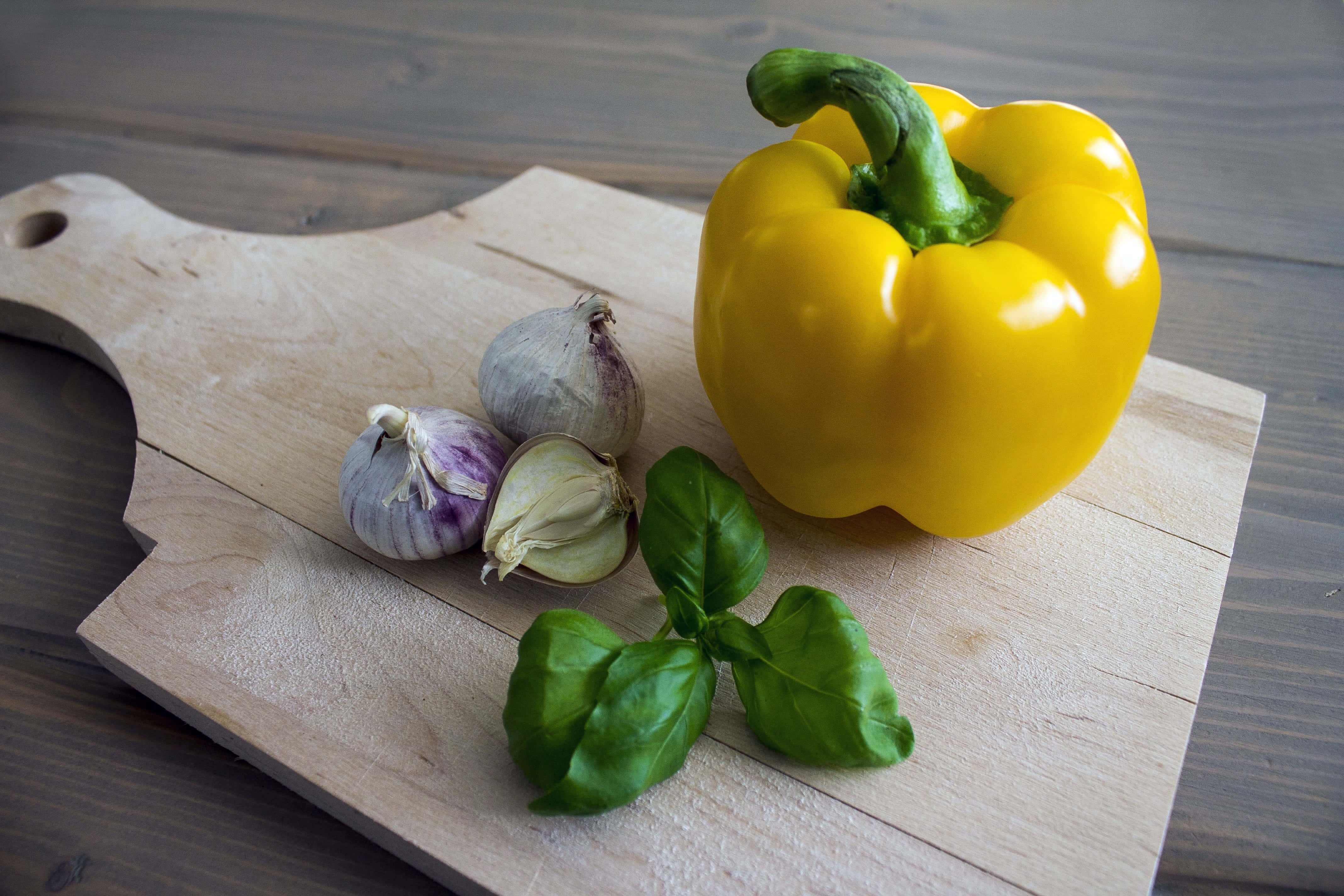 Yellow Bell Pepper 桌布 壁紙 — 一個鮮黃色的燈籠椒、蒜瓣和新鮮羅勒葉放在淺色木砧板上。