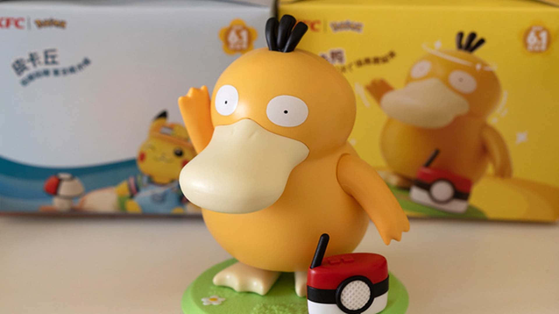 Pokemon Figures 桌布 壁紙 — 一個黃色卡通鴨子寶可夢公仔喺綠色底座上，喺精靈球旁邊開心咁揮手，後面有模糊嘅產品盒。