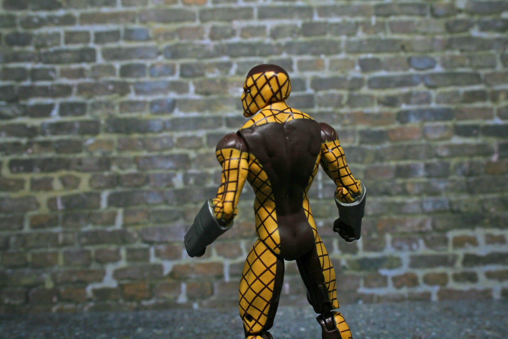 The Shocker 桌布 壁紙 — 一個黃色和棕色蜘蛛主題嘅動作人偶背向觀眾，企喺風化嘅磚牆前面。