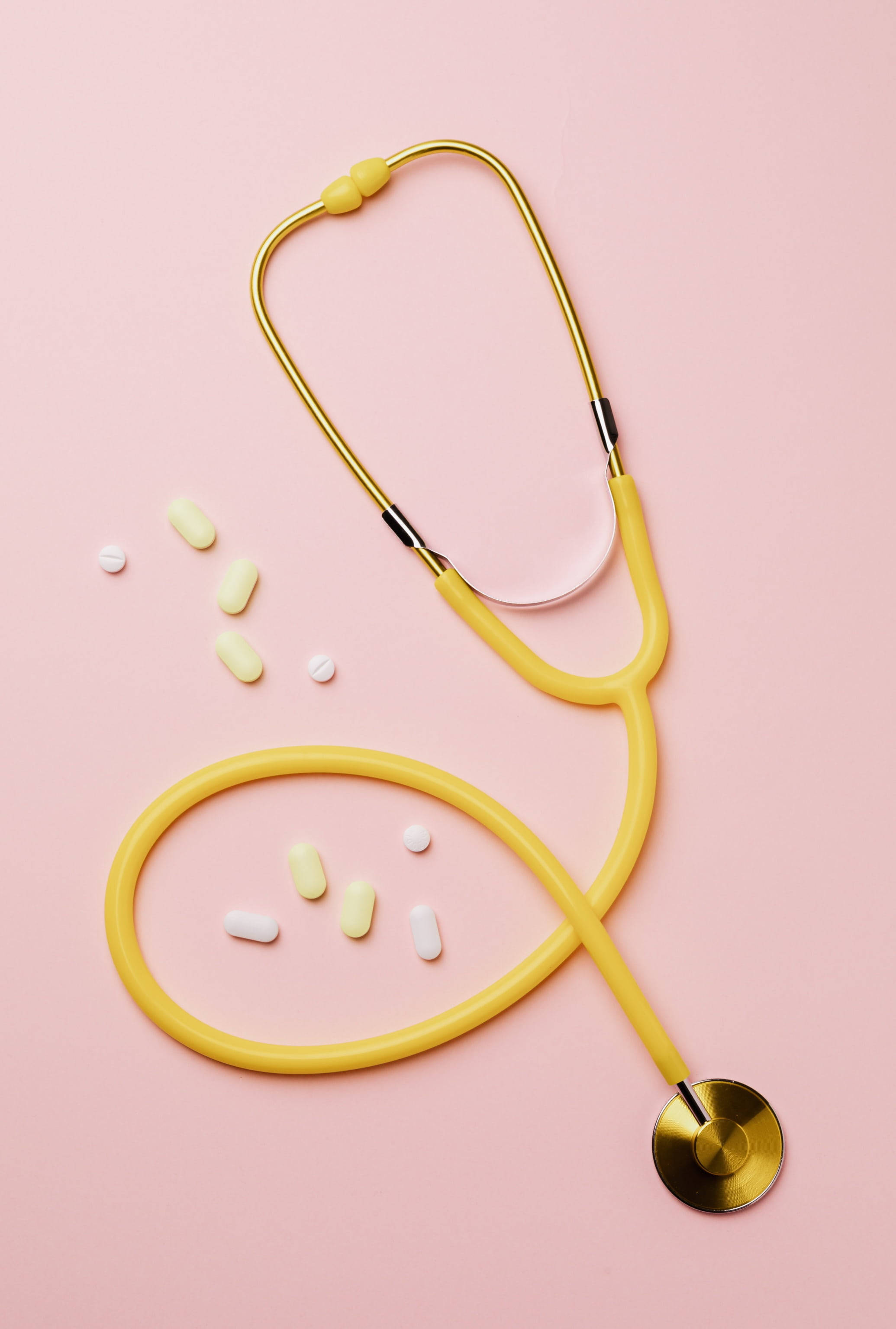 Stethoscope 桌布 壁紙 — 一個黃色聽診器和散落的藥丸放在柔軟的粉紅色背景上，形成極簡的平鋪佈局。
