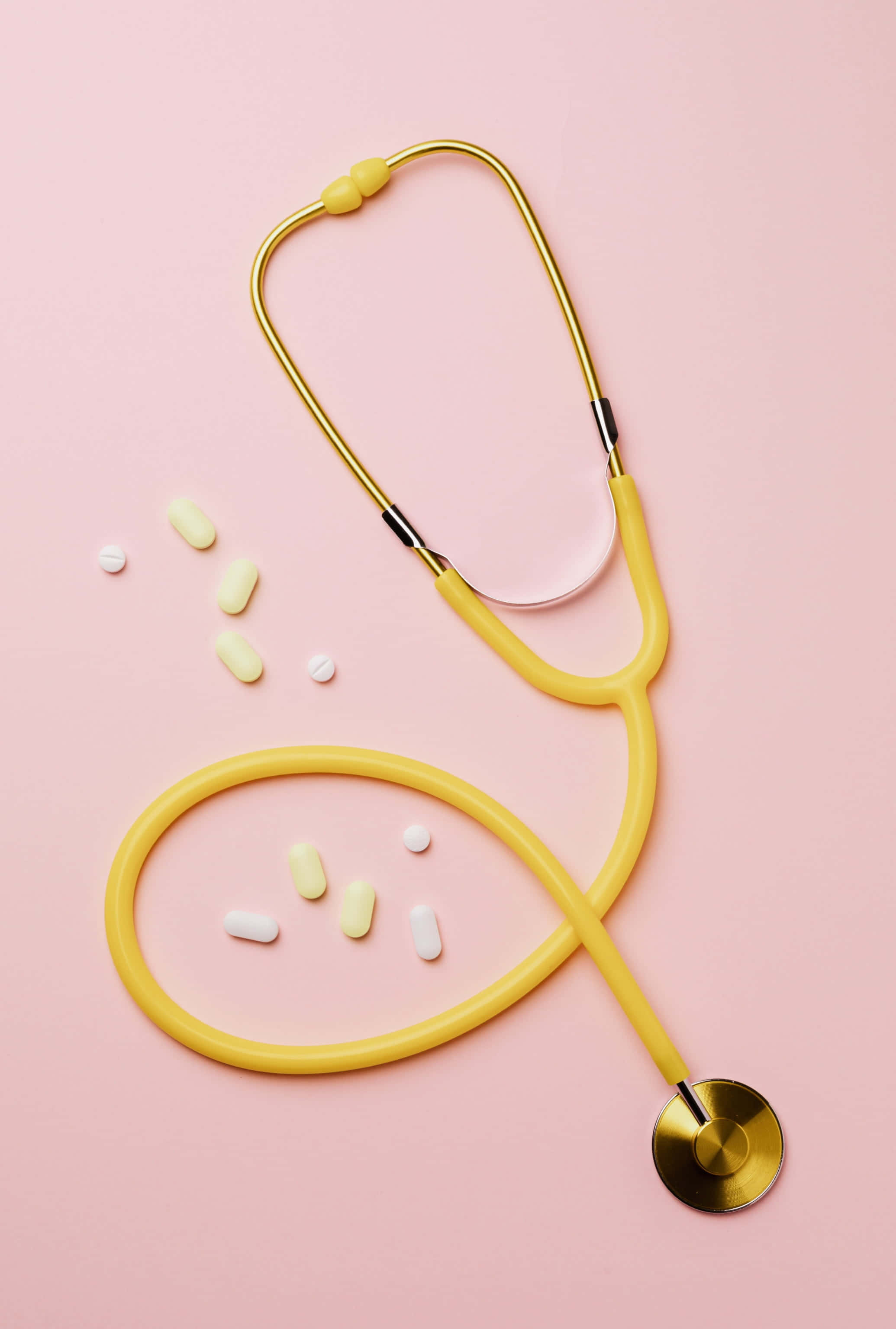 Stethoscope 桌布 壁紙 — 一個黃色聽診器和散落的藥丸放在柔軟的粉紅色背景上，呈現極簡的平鋪佈局。
