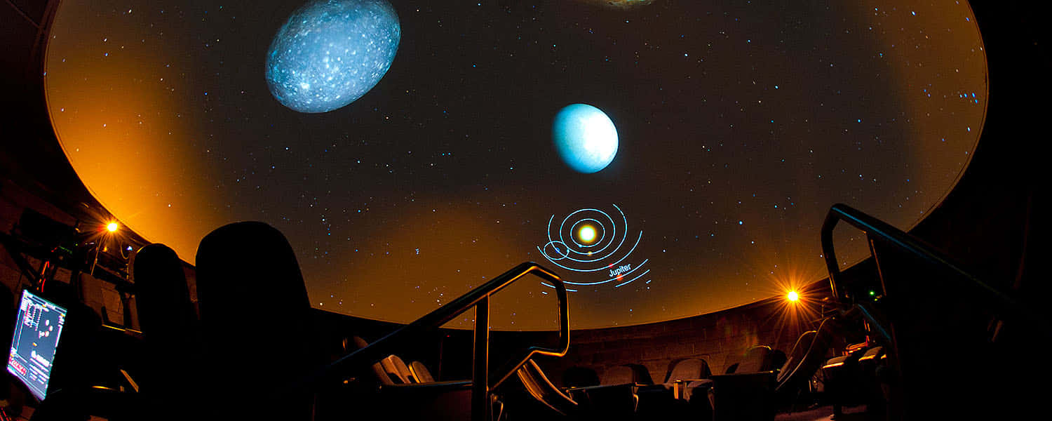 Planetarium 桌布 壁紙 — 一個黑暗的天文館穹頂顯示發光的藍色行星和星空，下方是一排排空座位。