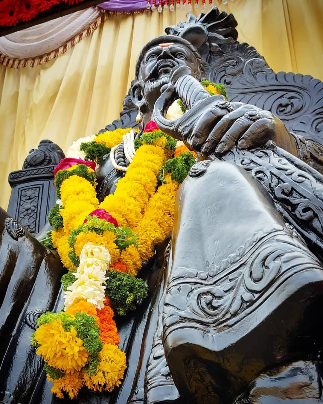 Chhatrapati Shivaji Maharaj 桌布 壁紙 — 一個黑暗華麗雕像的特寫，雕像上裝飾著鮮豔的黃色、橙色和白色花環，背景是垂墜的金色窗簾。