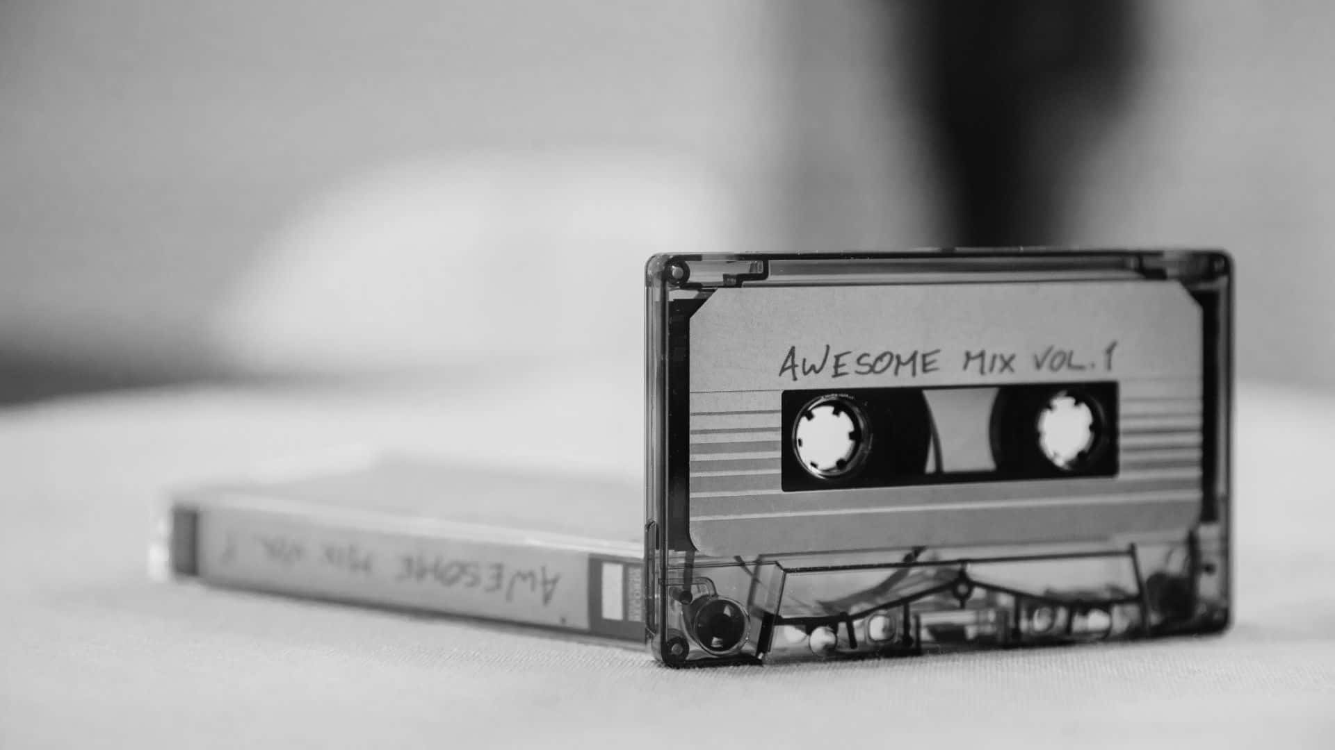 Black And White Music 桌布 壁紙 — 一個黑白特寫，透明卡式錄音帶標有「Awesome Mix Vol. 1」，放在柔軟表面上，旁邊有盒子。