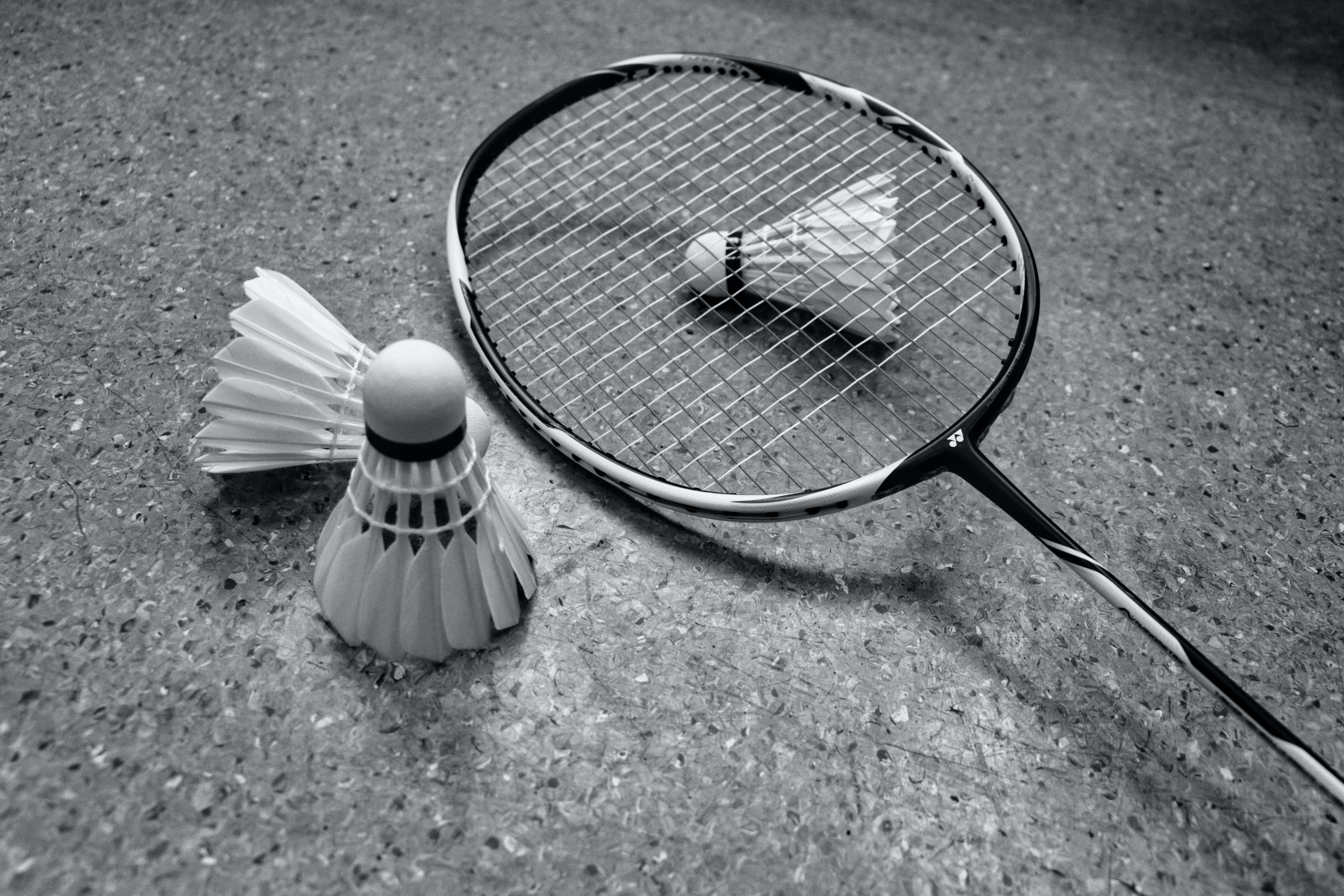 Badminton 桌布 壁紙 — 一個黑白特寫，顯示一個羽毛球拍和兩個羽毛球放在有紋理的路面上。